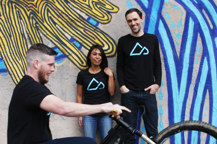 Three individuals are posing in front of a vibrant graffiti wall featuring blue and yellow patterns. The first person is doing a wheelie on a mountain bike and appears to be having fun. The second person, standing in the middle, is looking at the cyclist with a smile, while the third person on the right is also smiling. All three are wearing matching black t-shirts with a blue logo.