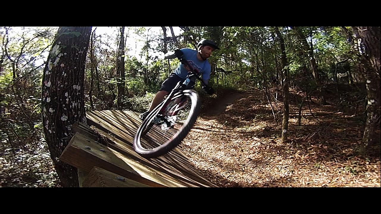 A mountain biker navigating a wooden ramp in a forested area, with trees and fallen leaves surrounding the trail. The rider is captured mid-maneuver, leaning into the turn with one wheel elevated off the ramp. Santos mountain bike trail.