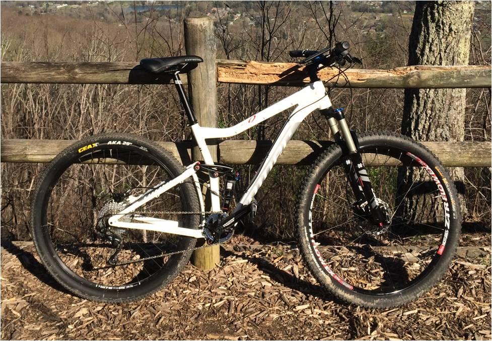 Niner Jet 9: Alt text: A white mountain bike is positioned next to a wooden fence in a wooded area. In the background, tree branches and a distant landscape are visible, suggesting an outdoor setting. The bike features thick tires and a suspension fork, indicating it is designed for off-road use.