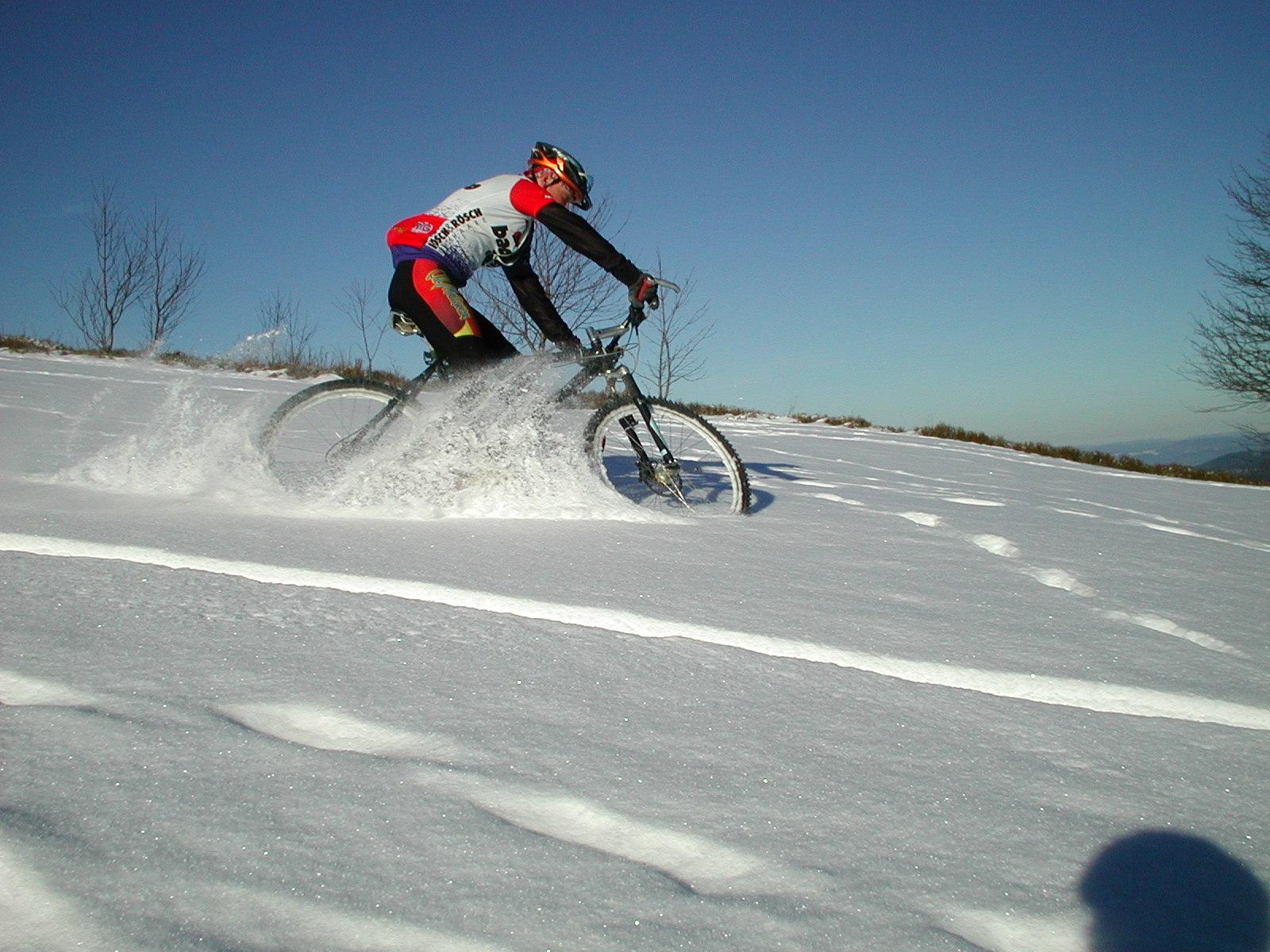 A person cycling on a mountain bike through a snowy landscape, leaving tracks in the fresh snow. The cyclist is wearing a helmet and a colorful cycling outfit, while the sky is clear and blue in the background. Sparse trees are visible on the horizon. Romero Road mountain bike trail.