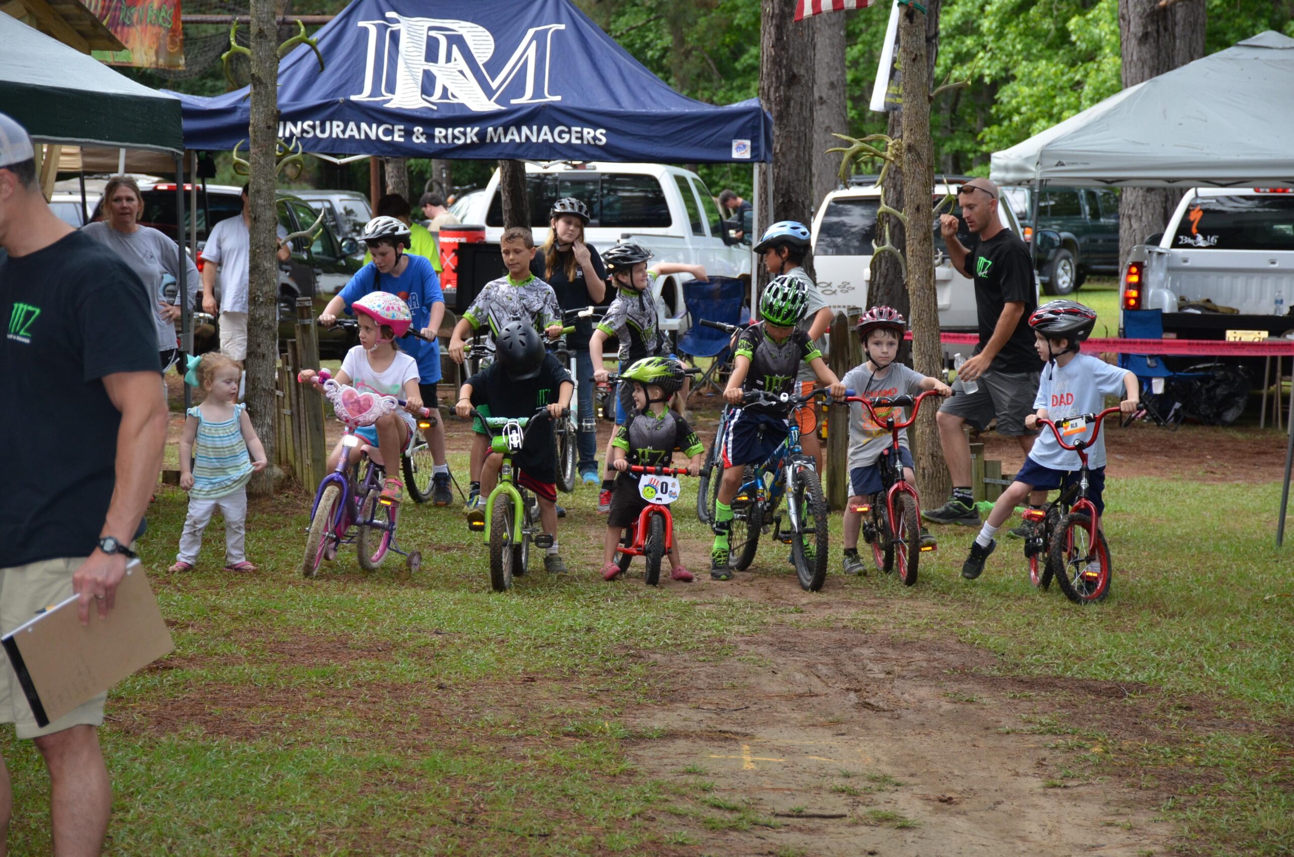 A group of children, ranging in age from toddlers to pre-teens, are lined up on bicycles, preparing for a race in a grassy area. Some kids are wearing helmets, and a few have decorated bikes. In the background, there are tents set up, and adults are watching or participating in the event. The atmosphere is full of excitement and anticipation for the upcoming race. Mt. Zion Bike Trails mountain bike trail.