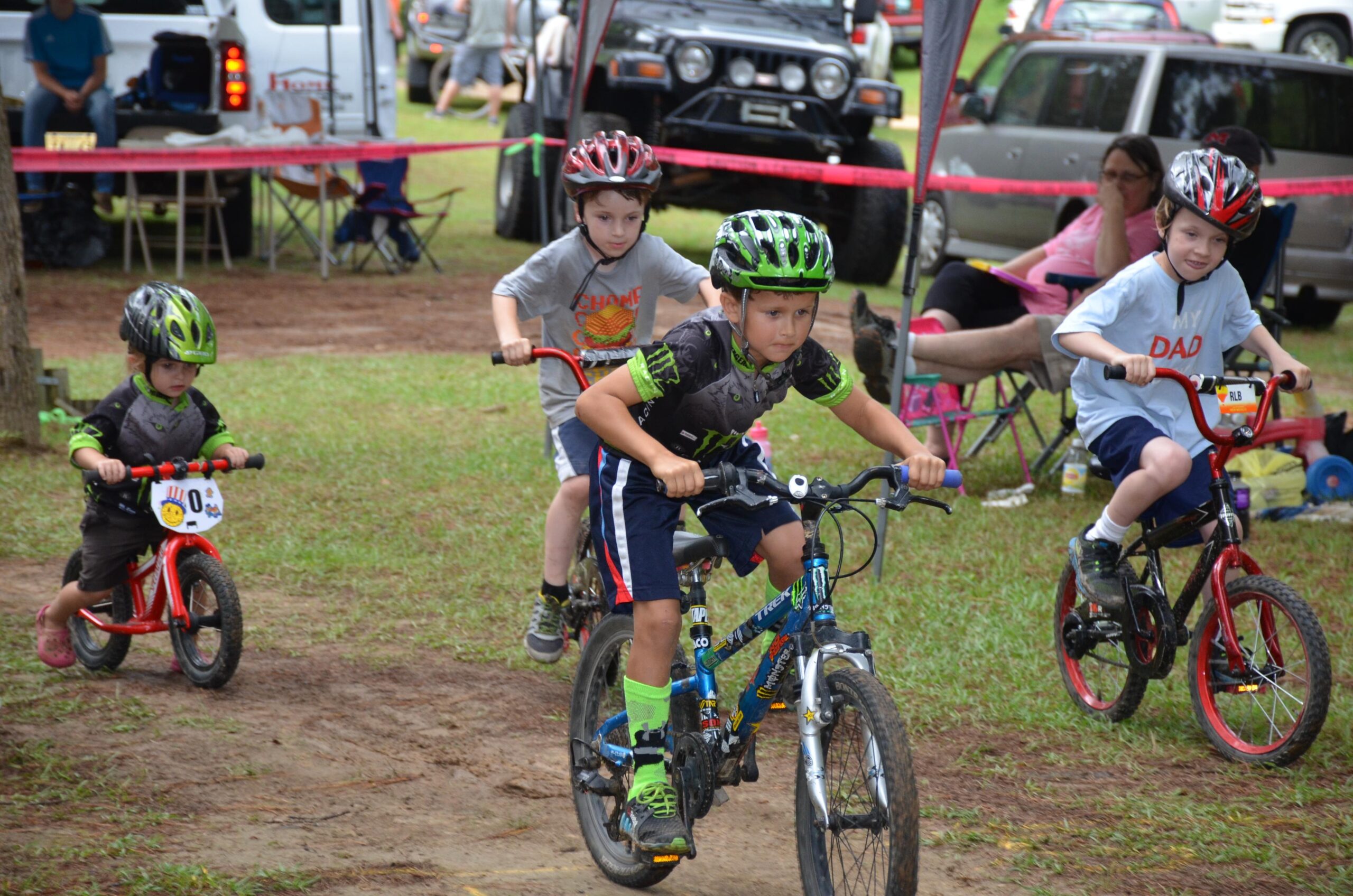 Four children in helmets ride bicycles along a dirt path in a grassy area, surrounded by parked vehicles. One child in the front is pedaling hard, wearing a green and black jersey. The others follow closely, each wearing helmets and varying clothing styles. A few adults can be seen in the background, observing and seated in foldable chairs. Mt. Zion Bike Trails mountain bike trail.