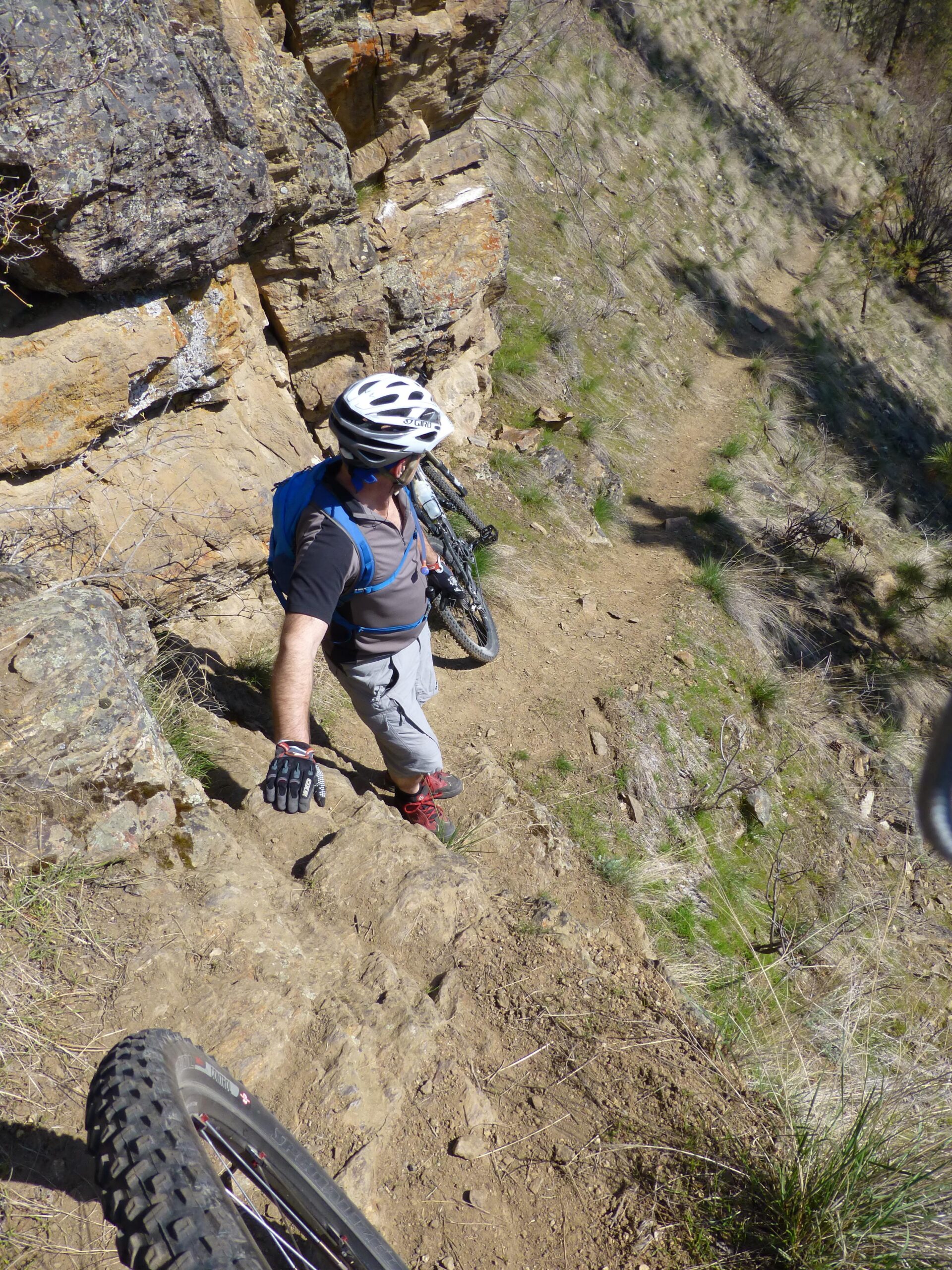 A person wearing a helmet and gloves is navigating a rocky, narrow path on a mountain biking trail. The individual is looking down the slope, with a bicycle resting nearby on the ground. The surrounding terrain features grass and scattered rocks, set against a backdrop of steep cliffs. The scene suggests an adventurous outdoor activity in a rugged landscape. Kalamalka Lake Provincial Park mountain bike trail.
