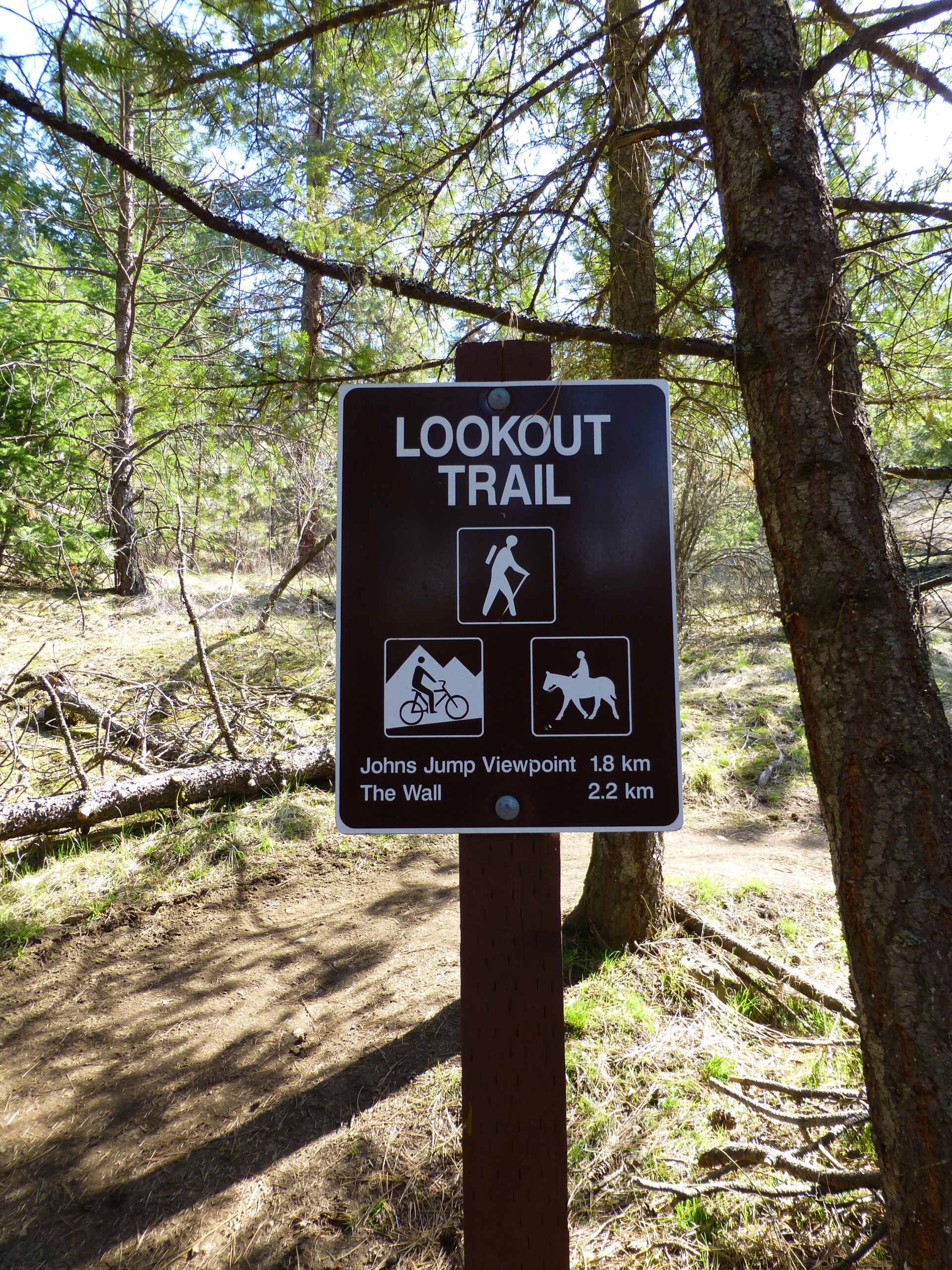 A trail sign reading "LOOKOUT TRAIL" with symbols indicating hiking, biking, and horseback riding. Directions to viewpoints "Johns Jump" at 1.8 km and "The Wall" at 2.2 km are also displayed. The background features trees and natural scenery. Kalamalka Lake Provincial Park mountain bike trail.