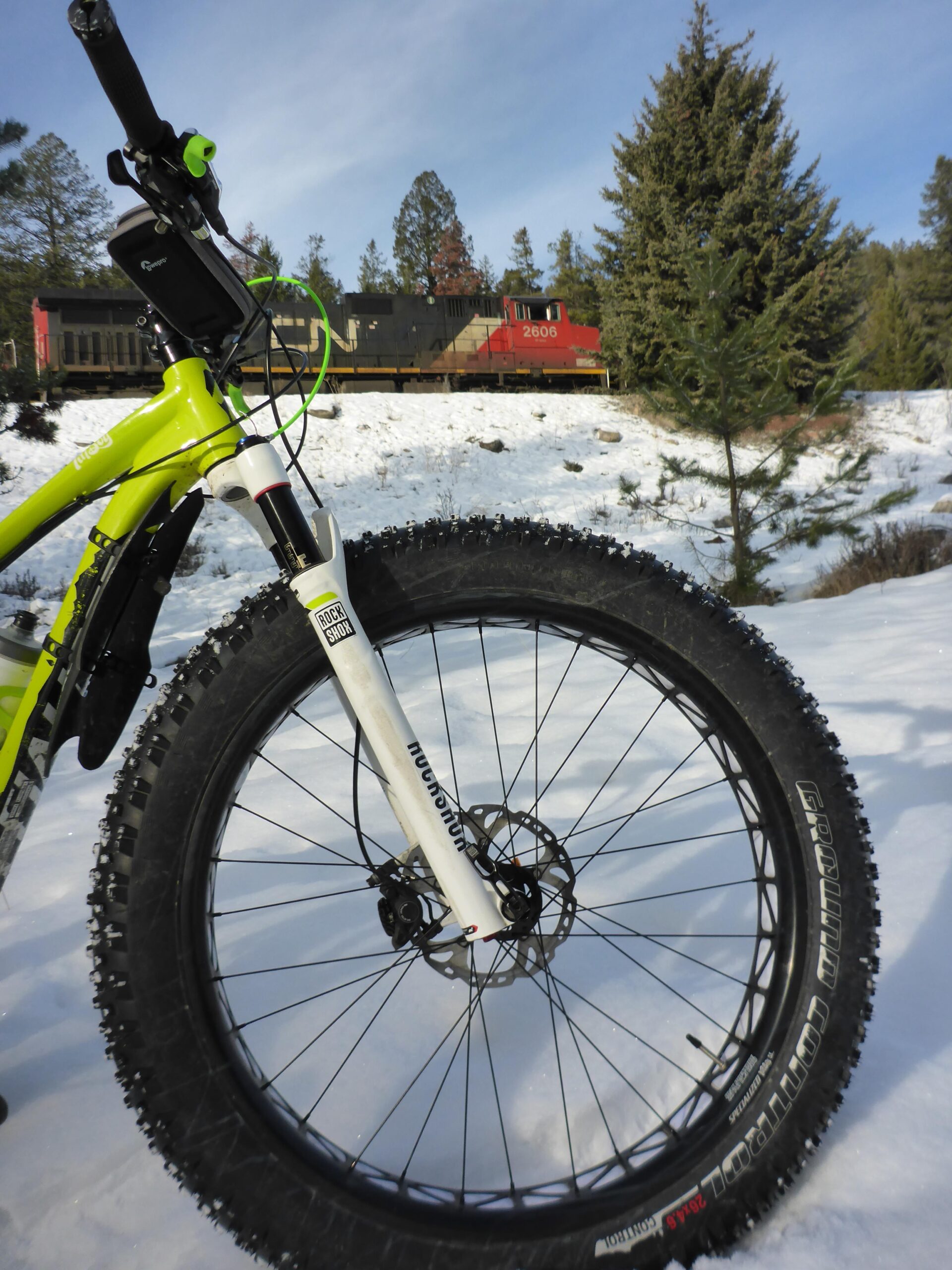 Specialized Fatboy Pro: A close-up view of a fat bike's front wheel and fork in a snowy landscape, with a train passing in the background. The bike is bright yellow, featuring thick tires designed for snow, and there are pine trees surrounding the scene.