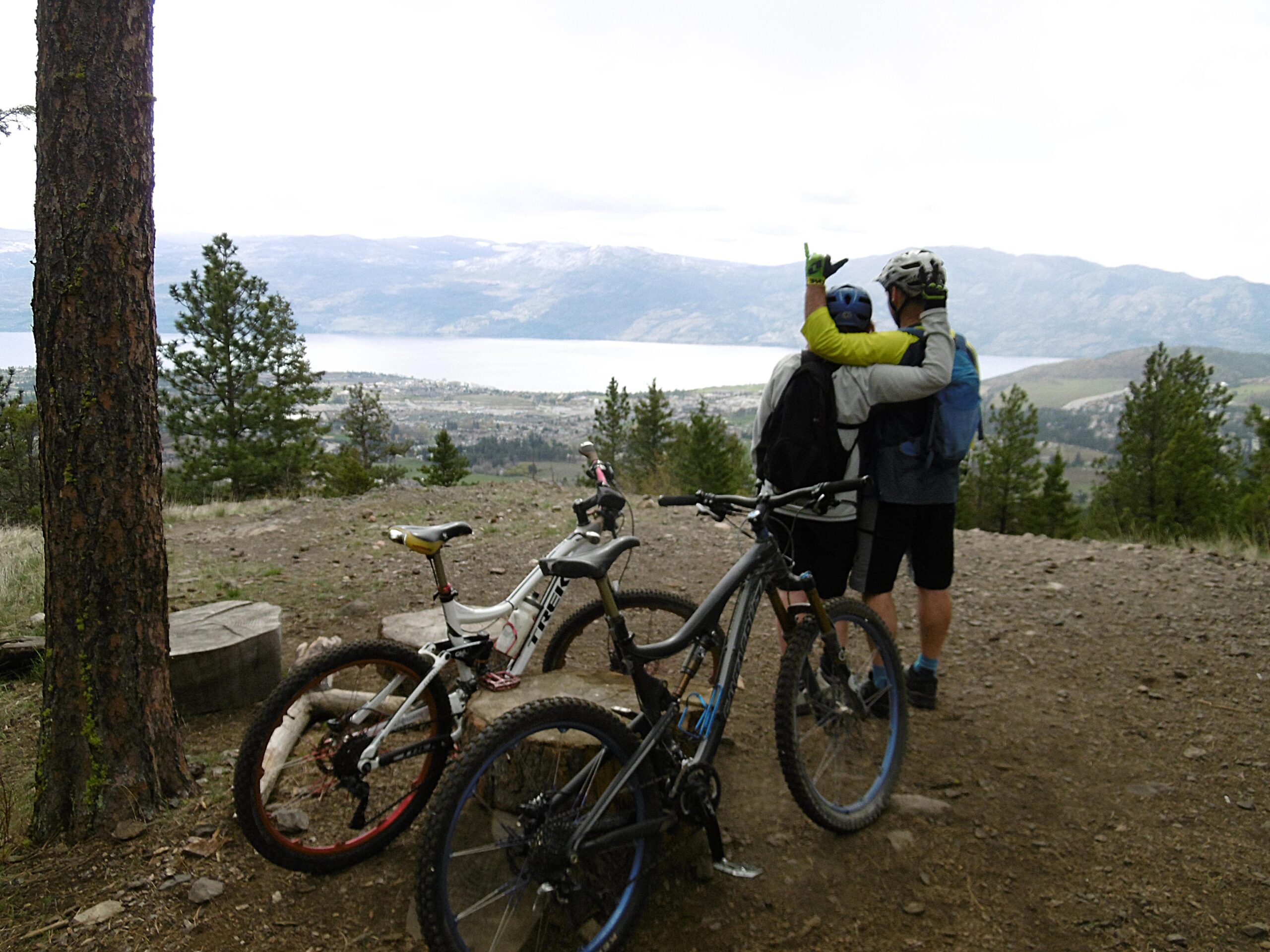 Two mountain bikers, wearing helmets and brightly colored apparel, embrace while overlooking a scenic landscape with a lake and mountains in the background. In the foreground, two bicycles are parked on a dirt trail surrounded by trees. Smith Creek mountain bike trail.