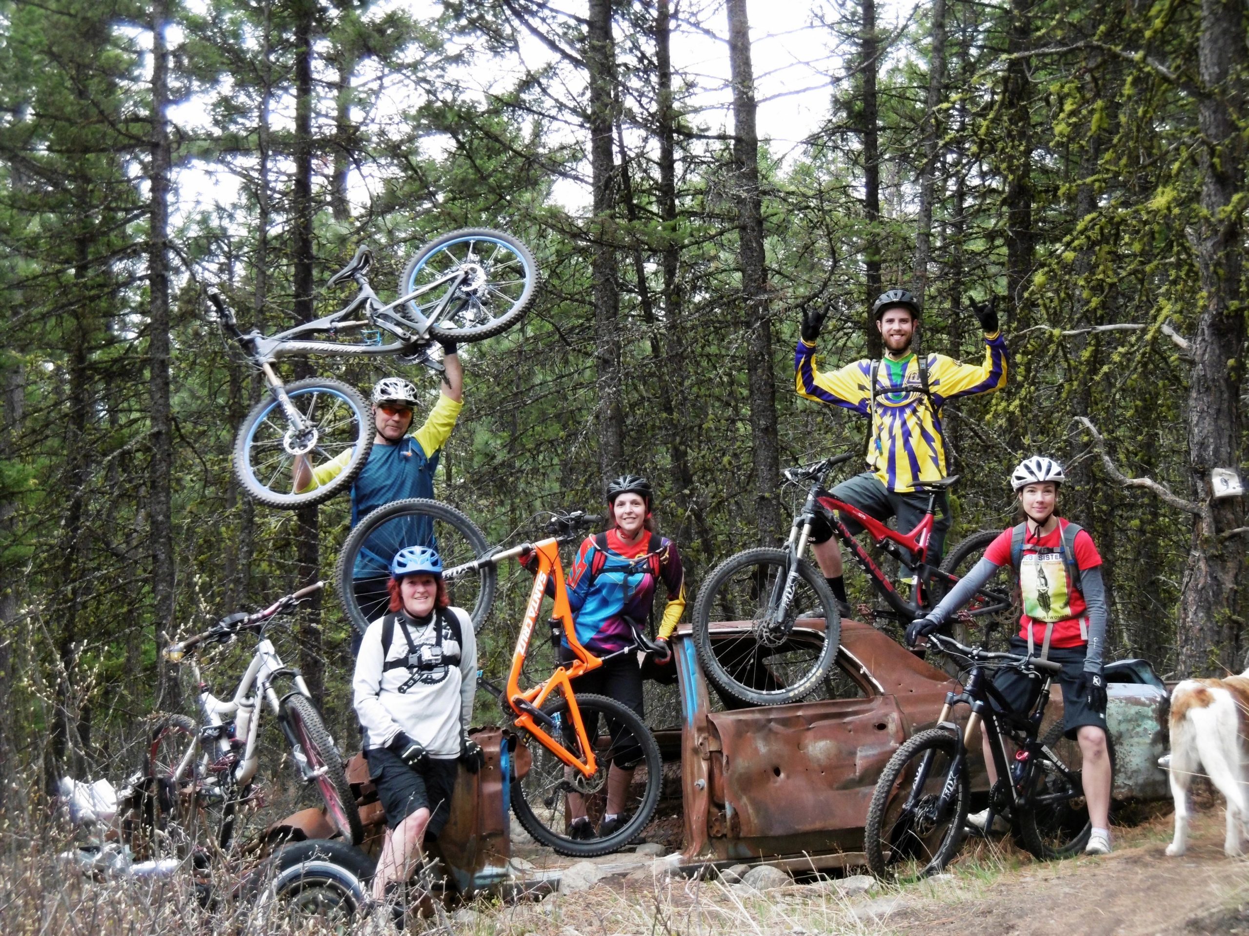 A group of five mountain bikers posing outdoors in a forest setting. They are standing on an old, rusty vehicle while holding their bikes. The group displays a mix of enthusiasm, with some members raising their hands in celebration. Surrounding them are tall trees, and a friendly dog is partially visible in the background. Smith Creek mountain bike trail.