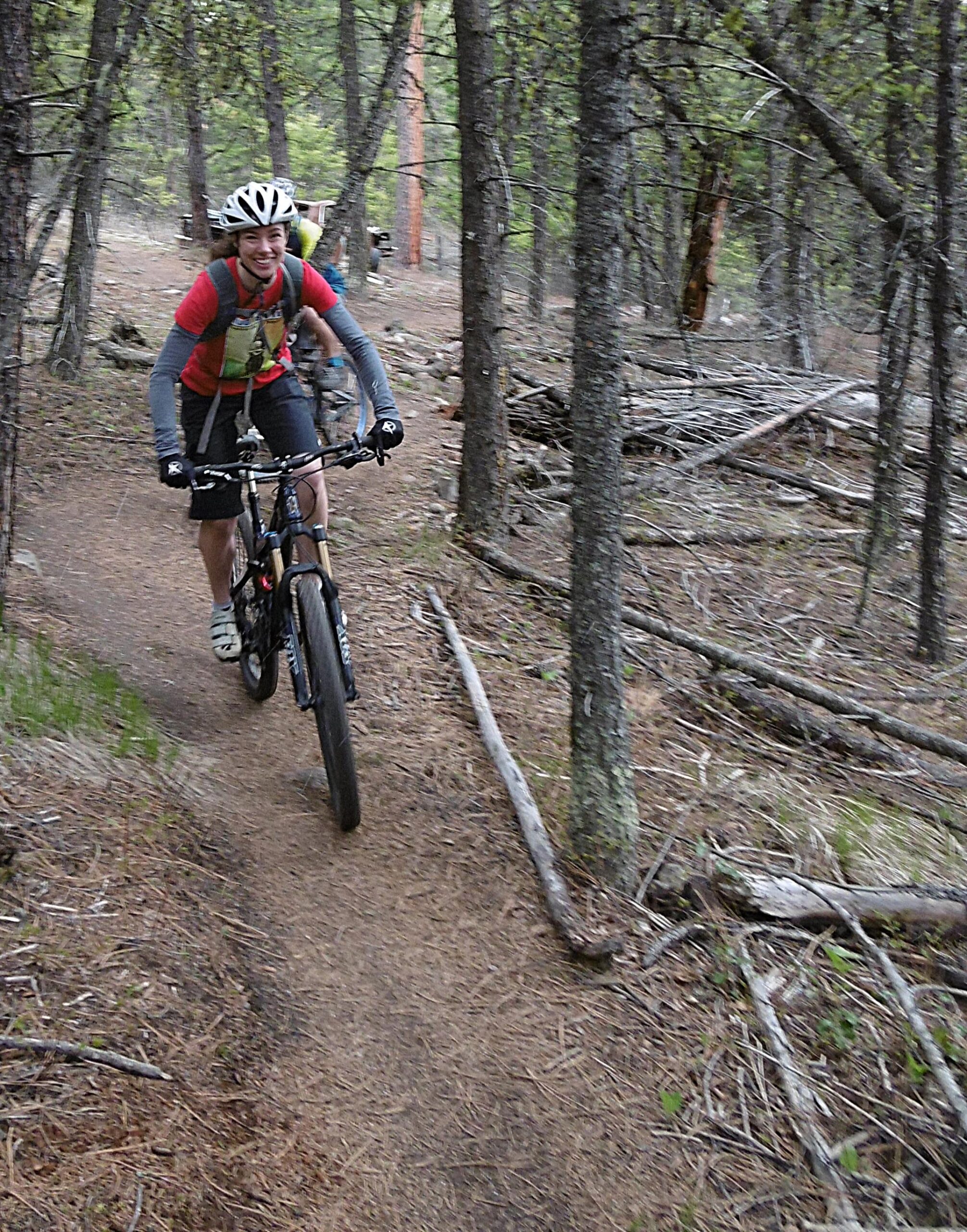 A cyclist riding a mountain bike on a dirt trail through a forested area, smiling and wearing a helmet and biking gear, with trees and fallen branches in the background. Smith Creek mountain bike trail.