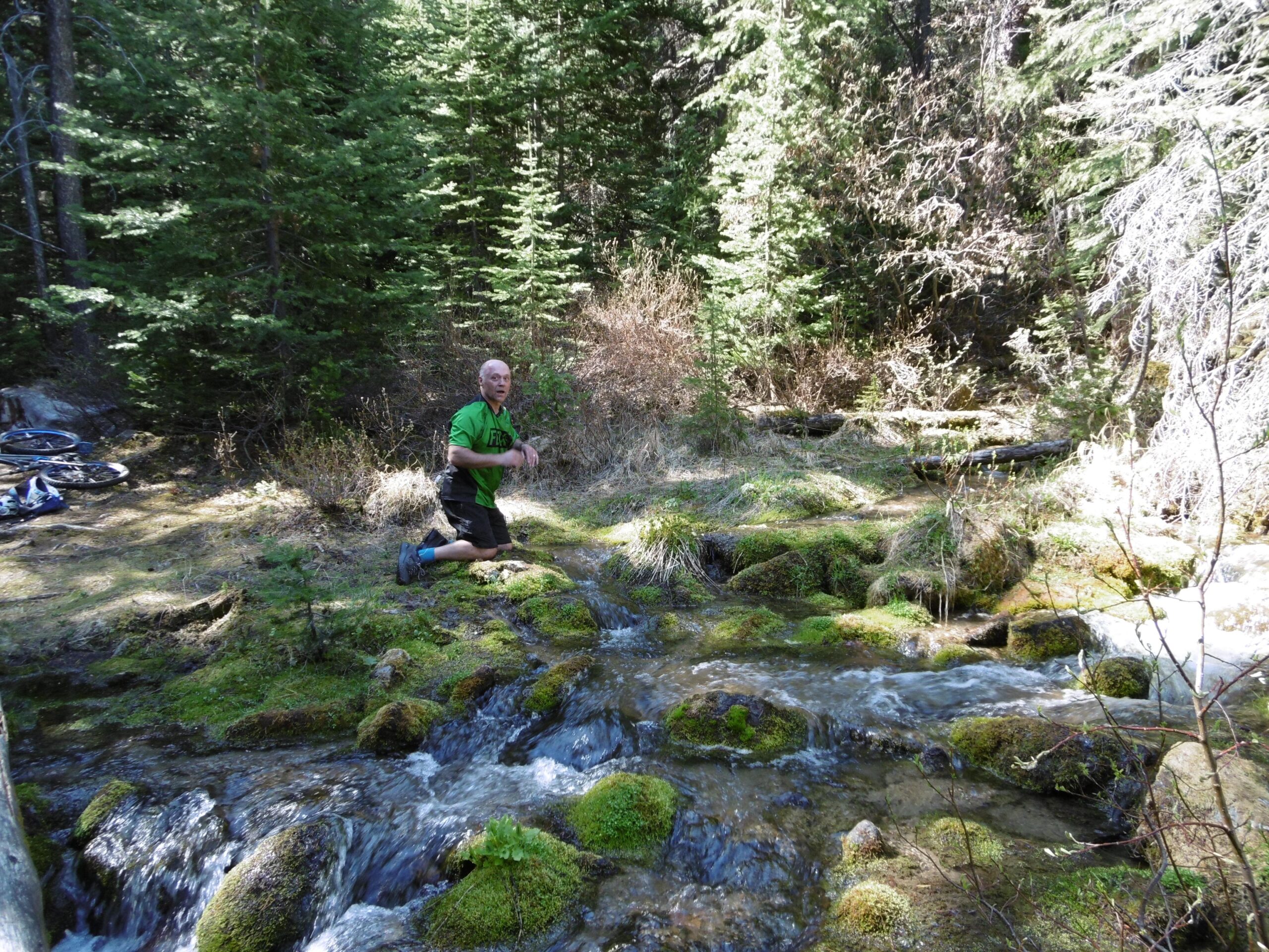 A person kneels by a flowing stream in a forested area, surrounded by lush greenery and moss-covered rocks. Mountain bikes are leaning against the trees in the background, indicating an outdoor adventure. Sunlight filters through the trees, creating a serene atmosphere. 20-mile loop / Saturday Night Lake Loop mountain bike trail.