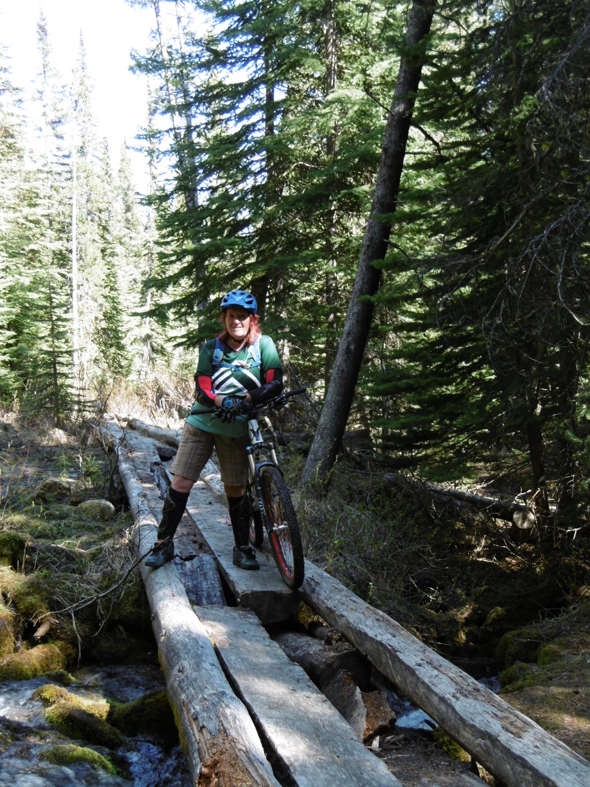 A person standing on a log bridge over a stream in a forested area, wearing a helmet, gloves, and biking gear. They are holding a mountain bike and smiling, with tall trees and bright blue sky in the background. The scene conveys an outdoor adventure in nature. 20-mile loop / Saturday Night Lake Loop mountain bike trail.