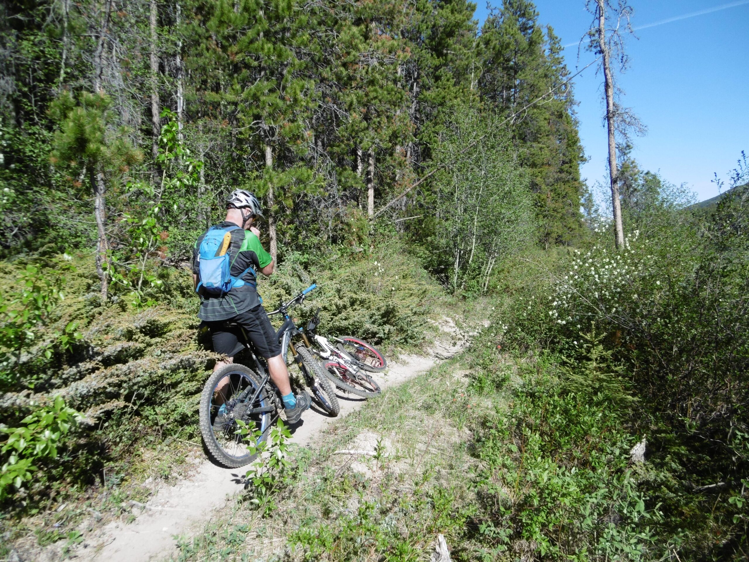 A mountain biker in a helmet and backpack stands next to his bike on a dirt path surrounded by lush greenery and trees. Another bike rests nearby as the rider appears to check a device or take a photo, enjoying a sunny day in nature. 20-mile loop / Saturday Night Lake Loop mountain bike trail.