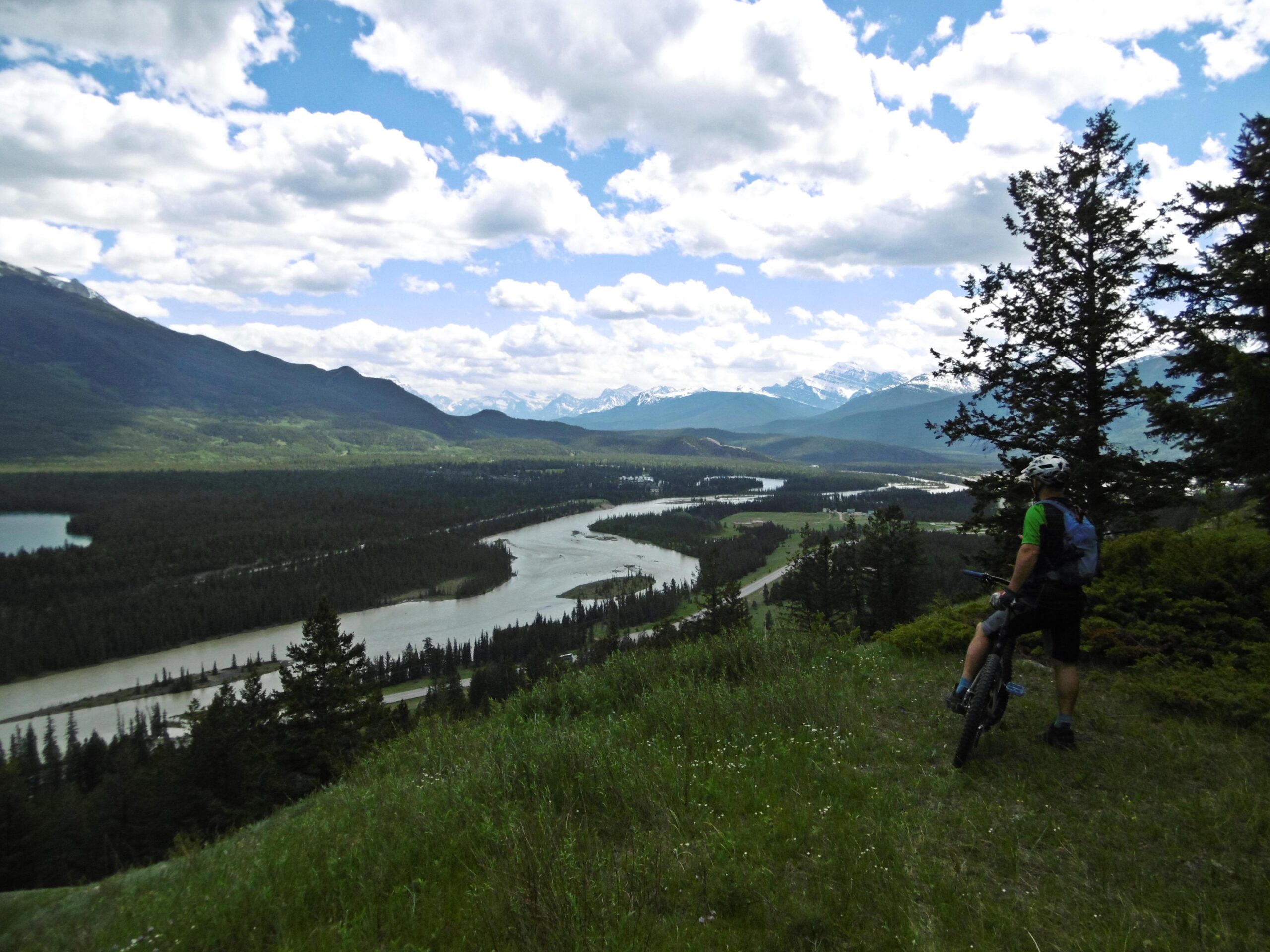 A mountain biker stands on a grassy hillside, looking out over a winding river surrounded by dense forests and mountains in the background. The sky is partly cloudy, adding depth to the vibrant scenery. Pyramid Bench mountain bike trail.