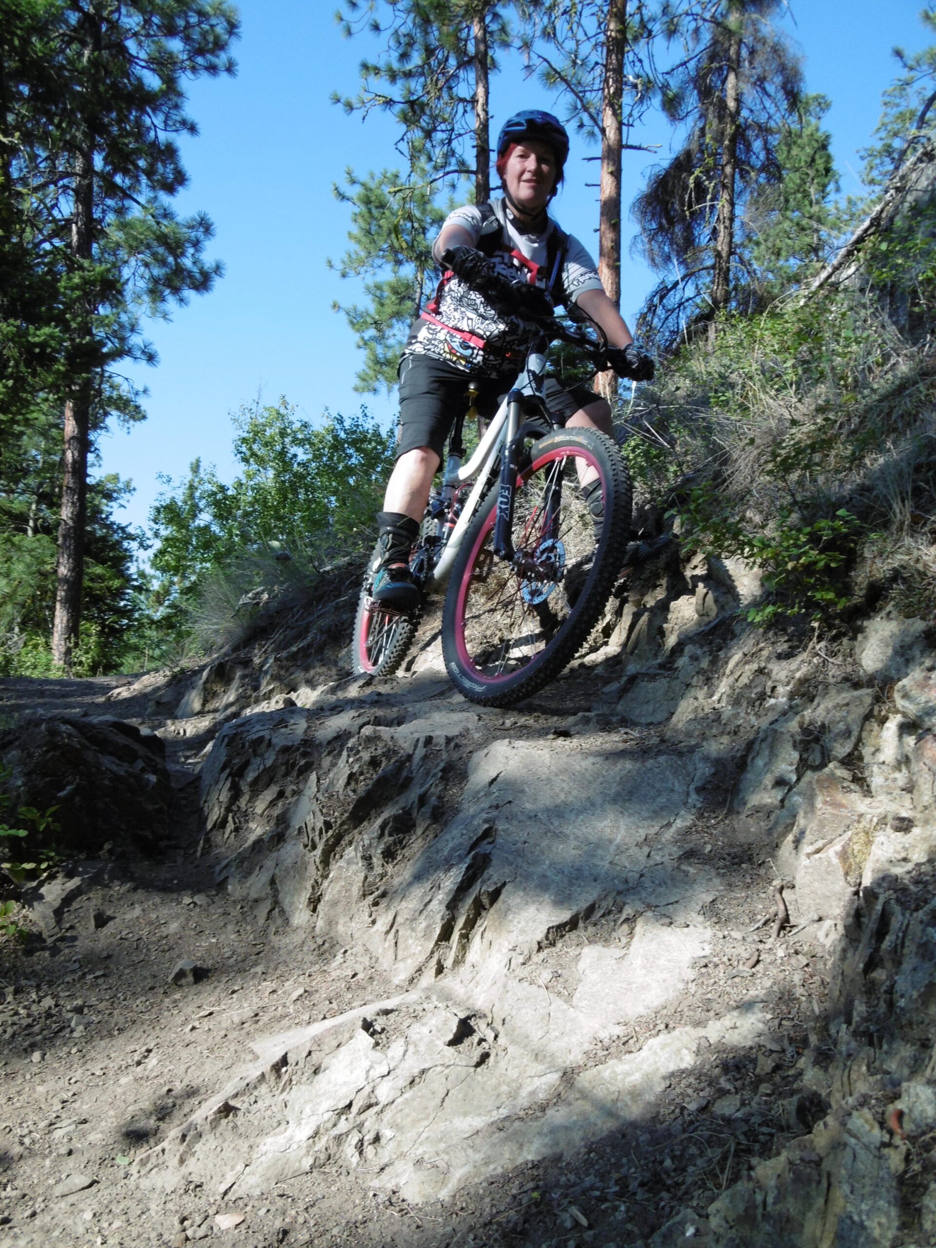 A mountain biker skillfully navigates a rocky trail surrounded by trees under a clear blue sky. The cyclist is wearing a helmet and riding gear, with a focus on maintaining balance while descending the rugged terrain. Rose Valley Resevoir mountain bike trail.