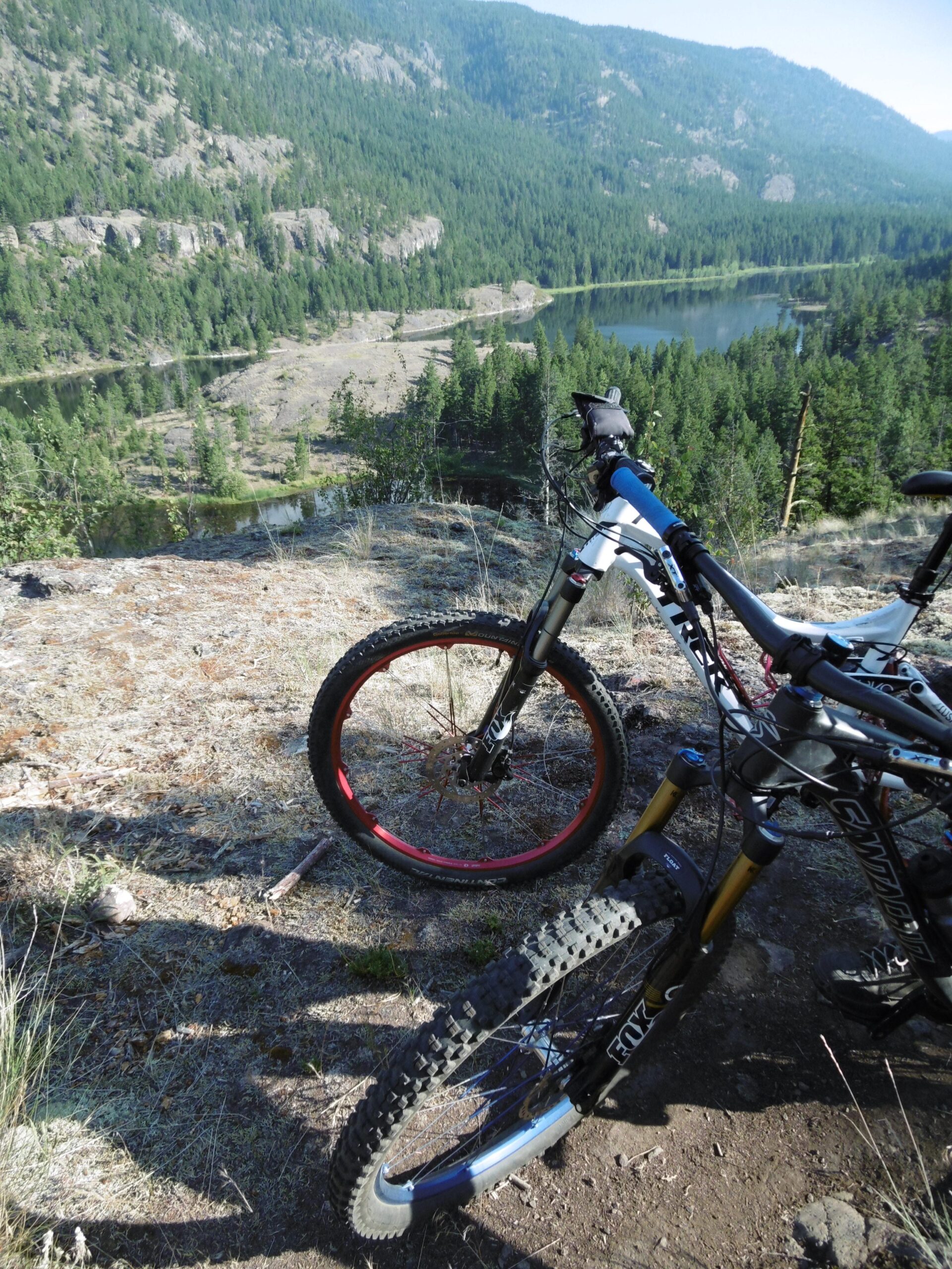 Two mountain bikes are parked on a rocky outcrop, overlooking a lush green landscape and a serene lake below. The background features dense forests and mountains, suggesting a scenic biking trail. The sunlight casts shadows of the bikes on the ground, highlighting the rugged terrain. Rose Valley Resevoir mountain bike trail.