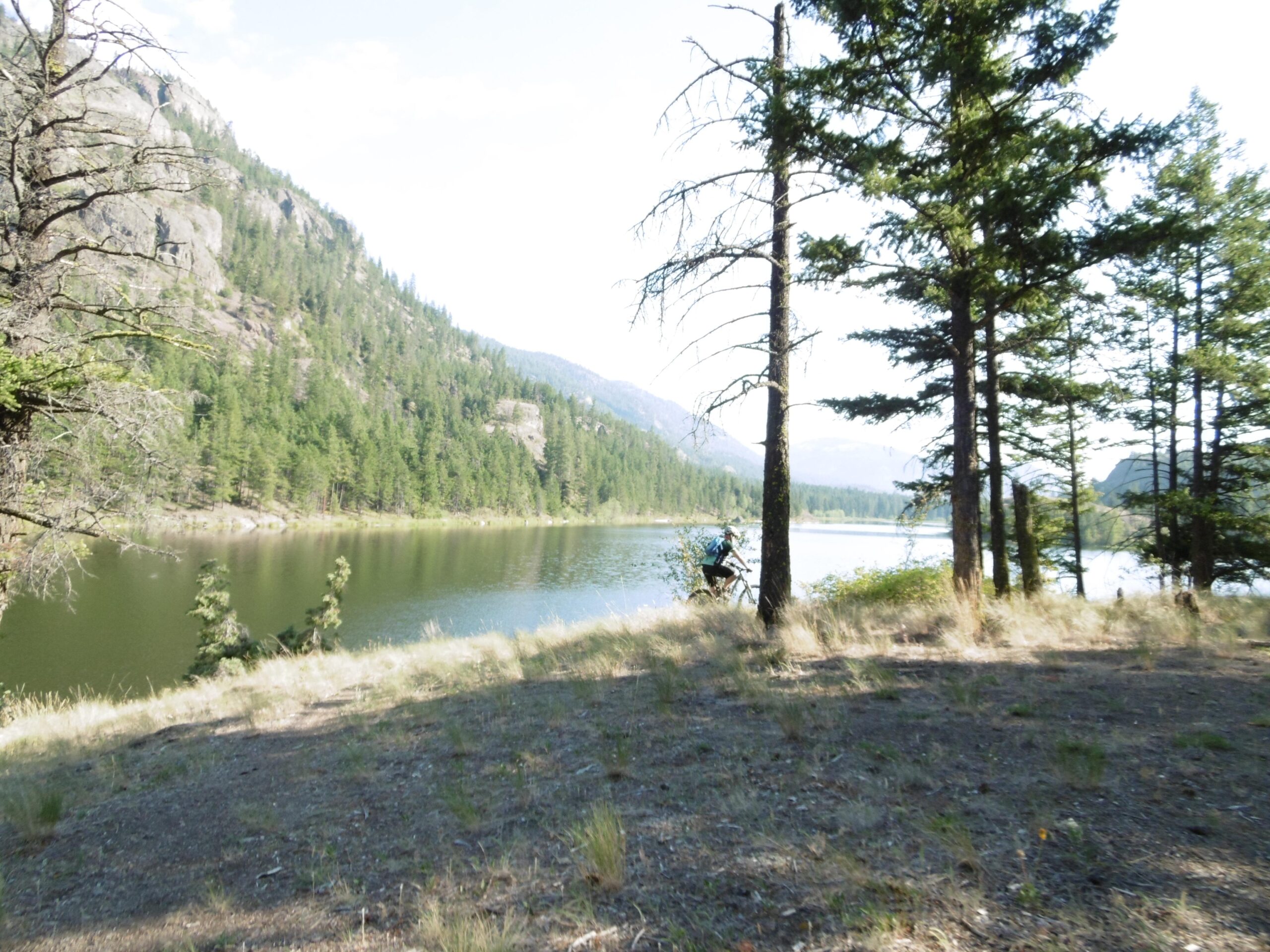 A serene landscape featuring a calm lake bordered by tall trees and mountain scenery in the background. The scene captures a peaceful natural environment with grassy shores and reflected trees in the water. Rose Valley Resevoir mountain bike trail.
