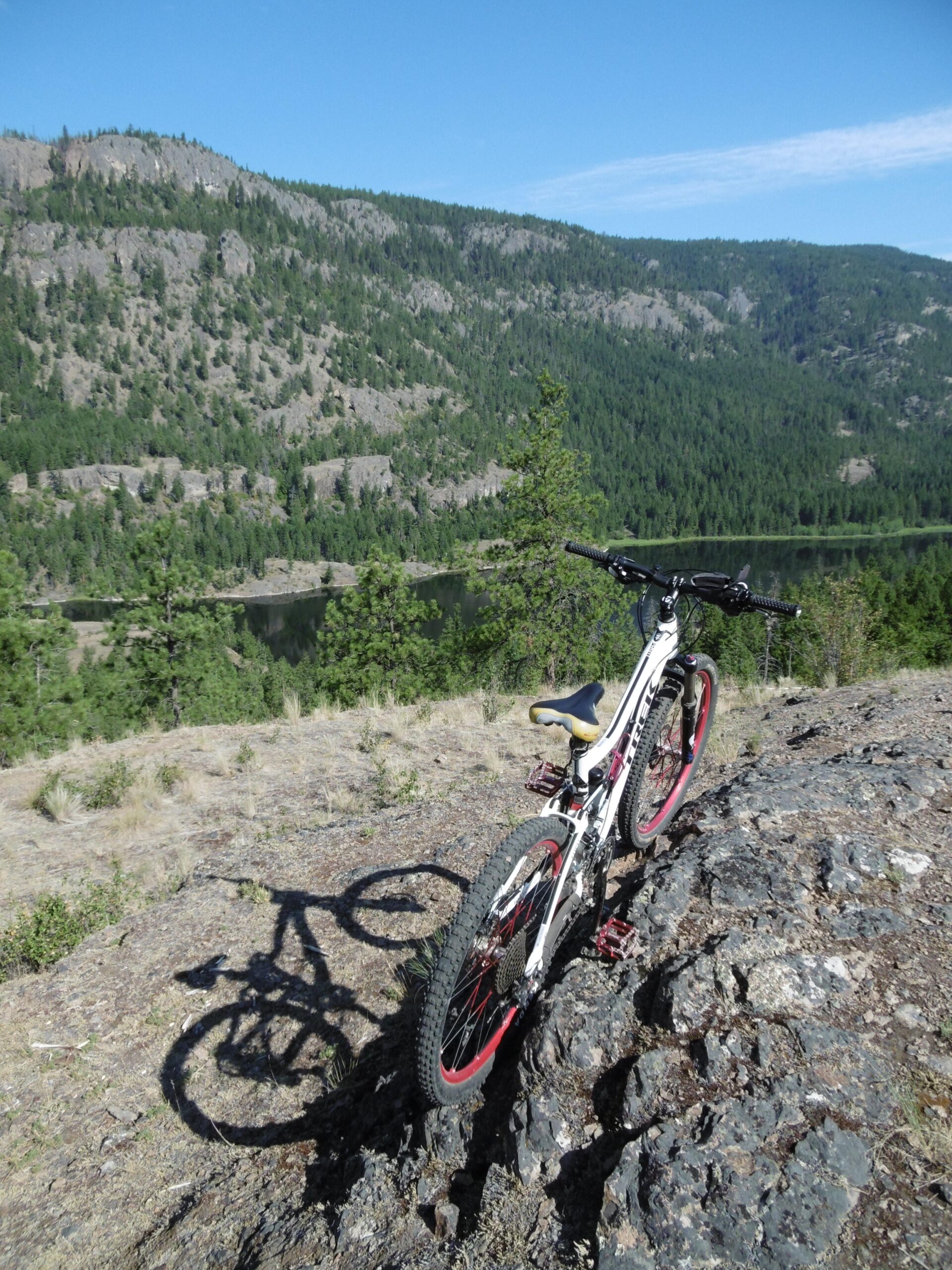 A mountain bike rests on a rocky outcrop overlooking a lush, green landscape with hills and a river below, under a clear blue sky. The bike casts a shadow on the rock surface, emphasizing its position in the natural scenery. Rose Valley Resevoir mountain bike trail.