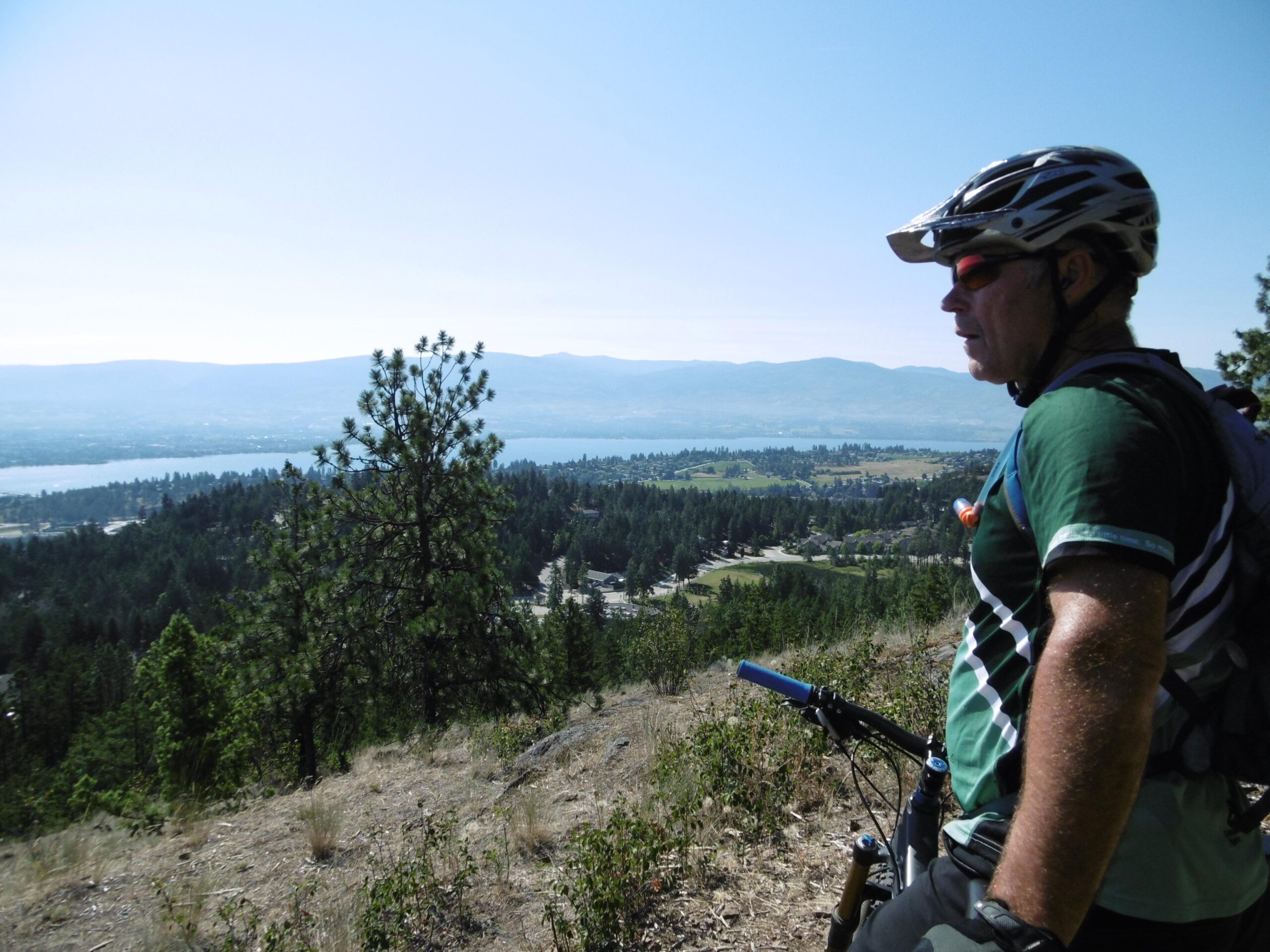 A mountain biker looking out over a scenic landscape, featuring a lake, rolling hills, and dense forests under a clear blue sky. The cyclist is wearing a helmet and sunglasses, with a focused expression, while standing beside a mountain bike. Rose Valley Resevoir mountain bike trail.