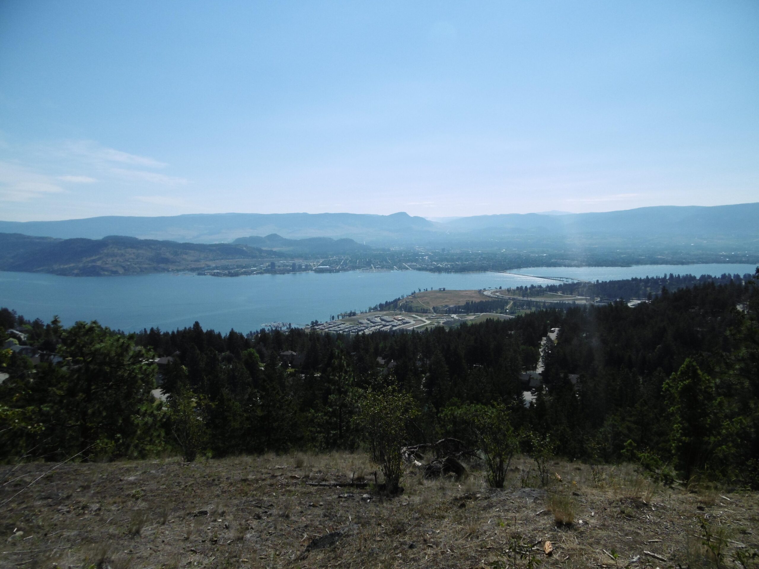 A panoramic view of a lake surrounded by hills and mountains on a clear day. In the foreground, there are patches of greenery and a few scattered trees, while the water reflects the blue sky. The shoreline features a small town with buildings and roads, alongside a marina with boats. The distant mountains create a scenic backdrop. Rose Valley Resevoir mountain bike trail.