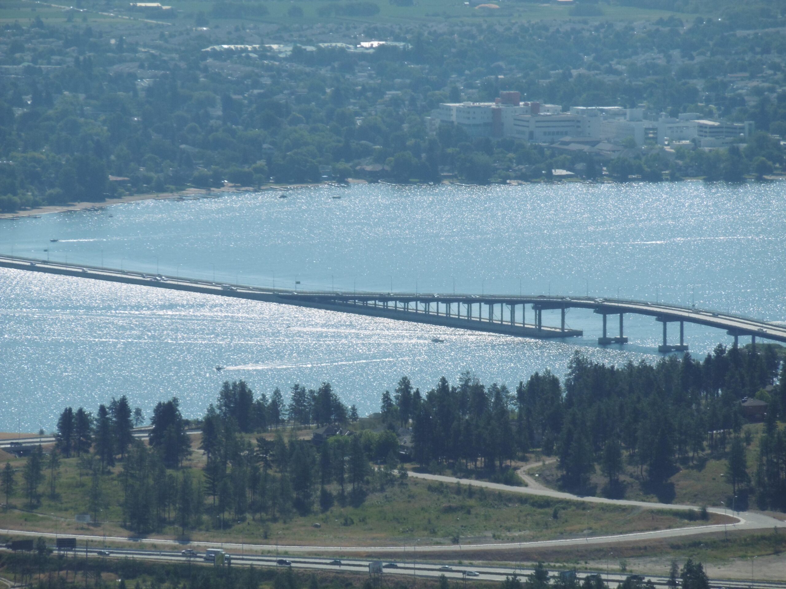 A scenic view of a long bridge spanning across a shimmering body of water, surrounded by lush greenery and a small town in the background. The sunlight reflects off the water's surface, creating a sparkling effect. Trees and winding roads are visible in the foreground, adding to the tranquil landscape. Rose Valley Resevoir mountain bike trail.