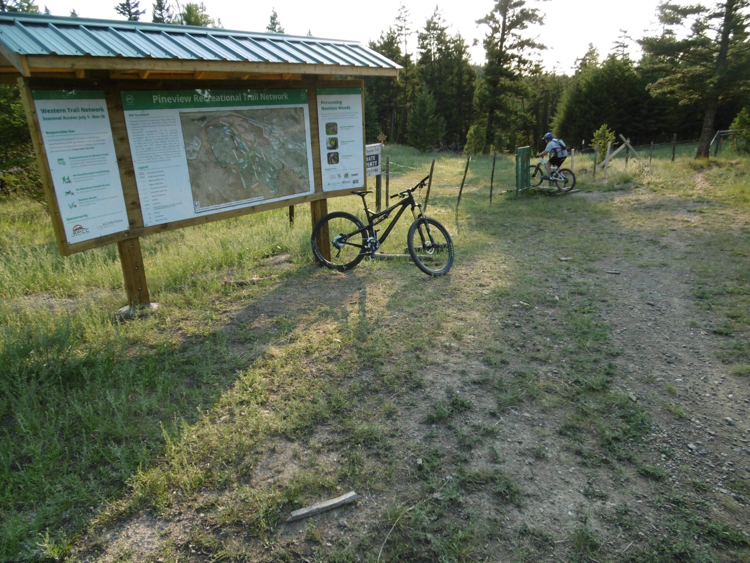 A scenic view of the Pineview Recreational Trail Network sign, displaying a map and information about the trails. In the foreground, a mountain bike is resting against the sign. In the background, a cyclist is seen approaching a gate, surrounded by lush green grass and trees. Pineview mountain bike trail.