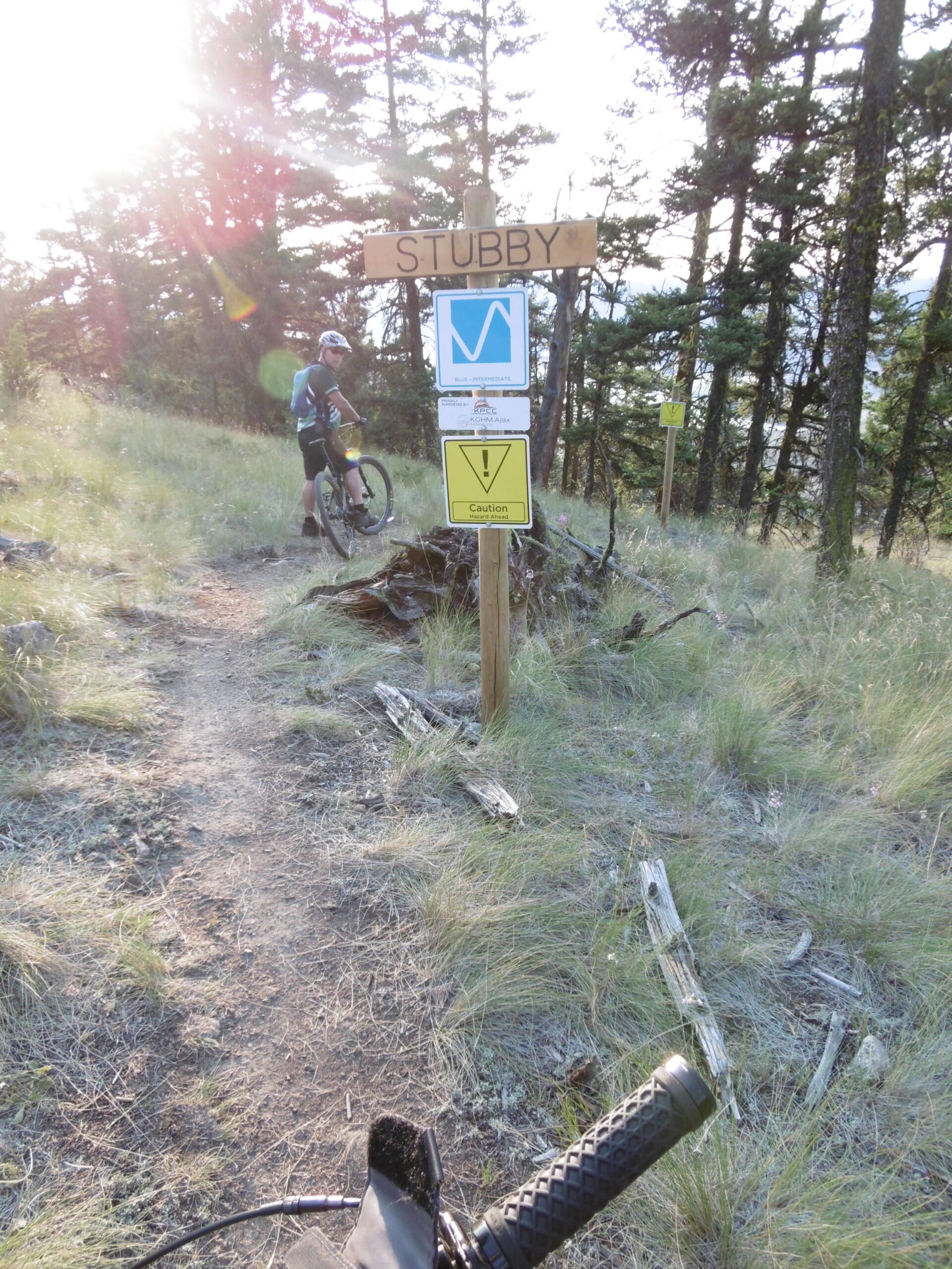 A mountain biker stands beside a wooden sign marked "STUBBY," indicating a blue intermediate trail. The sign includes additional caution signage and is surrounded by a lush, grassy area with trees in the background. The sun is shining, creating a bright atmosphere along the trail. Pineview mountain bike trail.