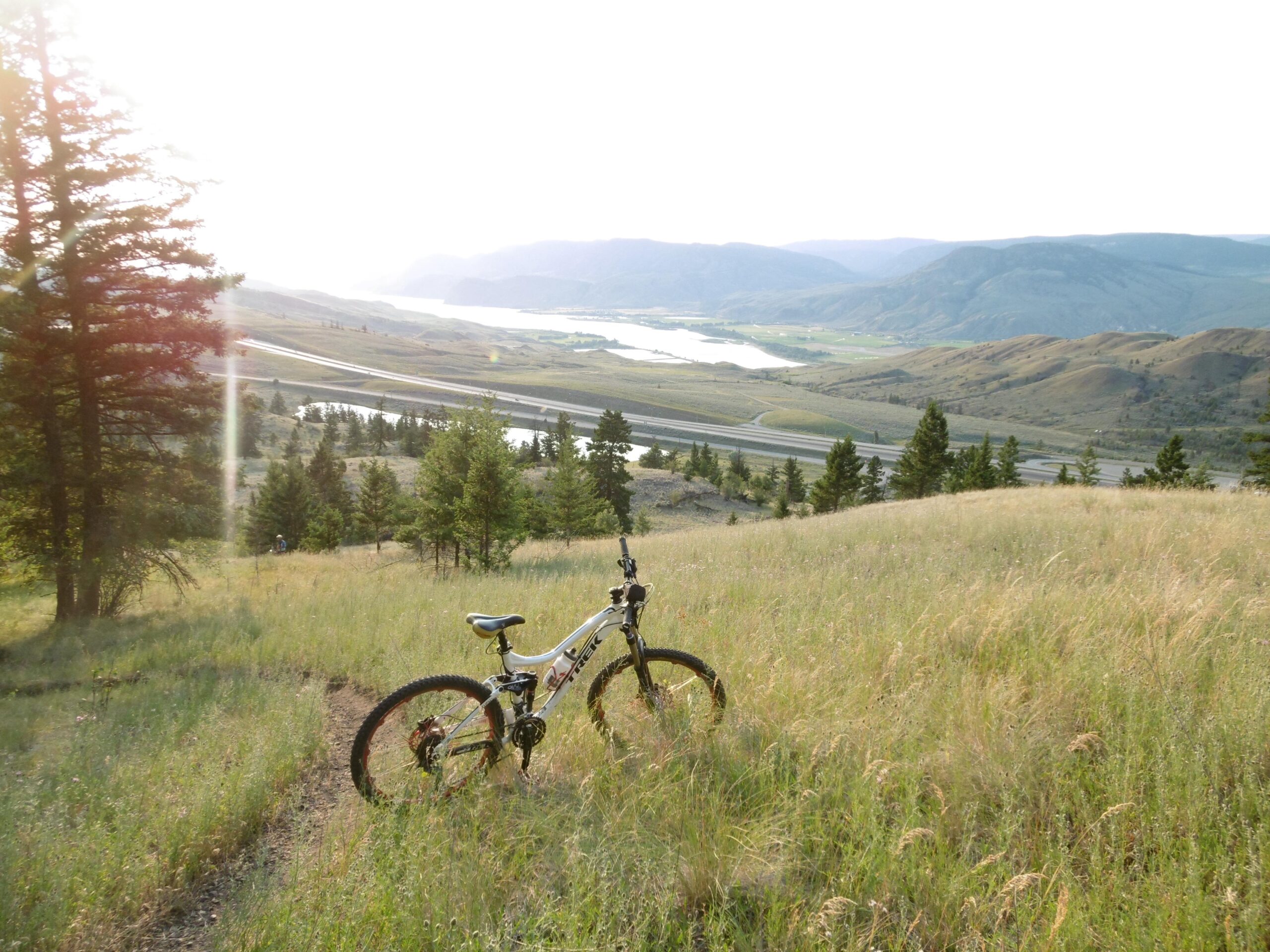 A mountain bike resting on a grassy trail overlooking a scenic valley at sunset. Rolling hills and a winding river are visible in the background, surrounded by trees and open fields. The scene is illuminated by warm, golden sunlight. Pineview mountain bike trail.