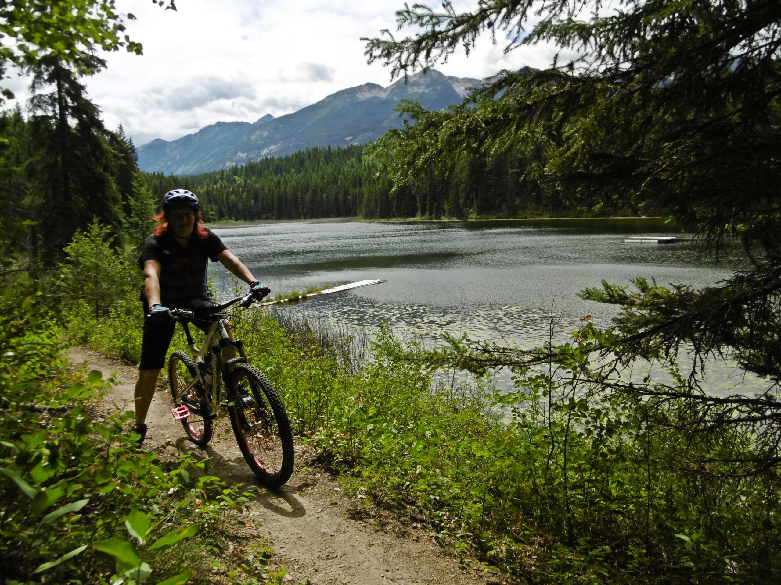 A person standing next to a mountain bike on a dirt path near a tranquil lake, surrounded by lush greenery and mountains in the background under a partly cloudy sky. Moonraker Trails mountain bike trail.