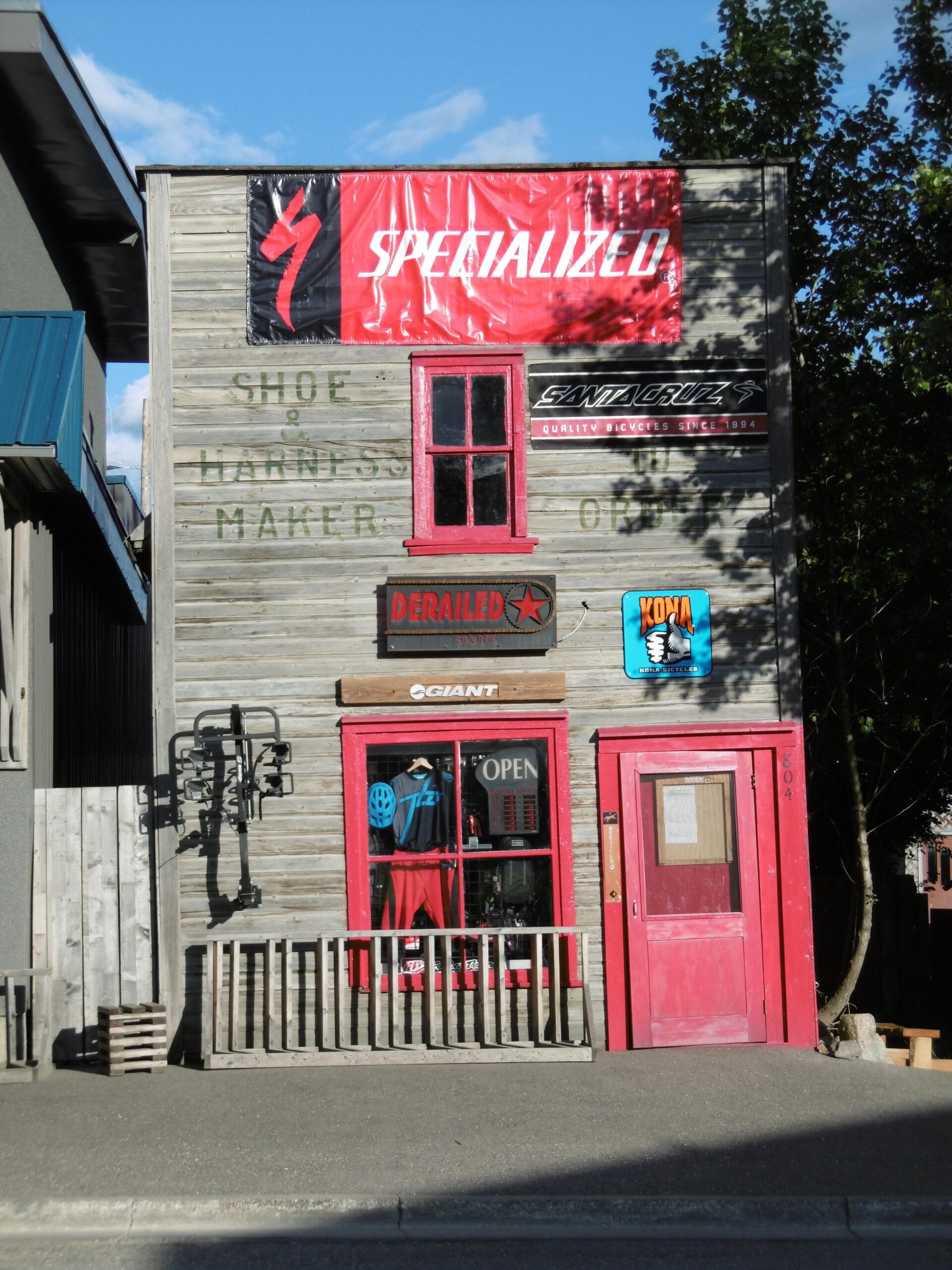 A rustic wooden building with a red door and window frames, featuring an "OPEN" sign. The facade displays various cycling-related banners and signage, including "Specialized," "Santa Cruz," "Giant," and "Derailed." The store