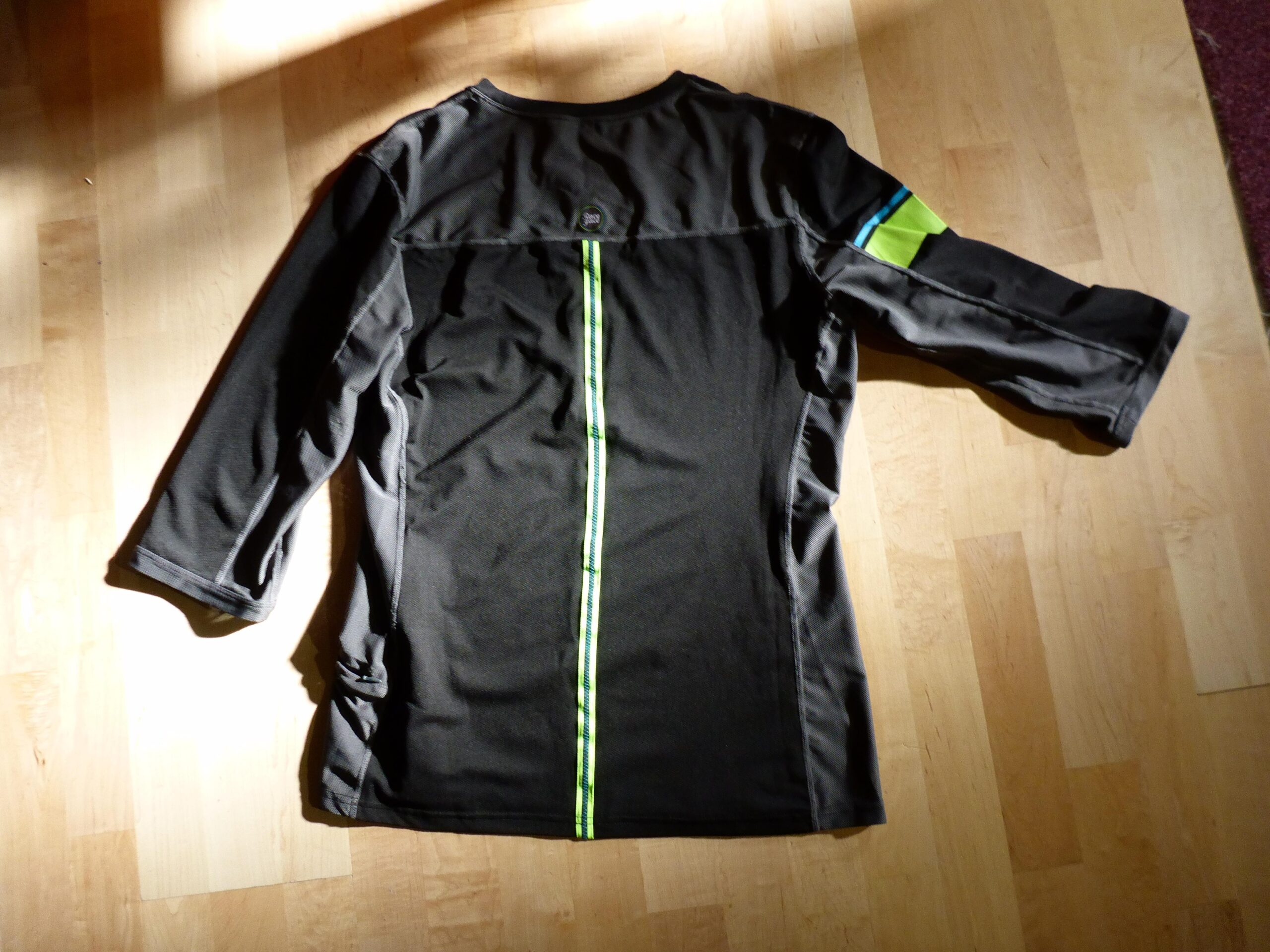 Race Face Khyber Jersey 3/4: A black long-sleeve shirt viewed from the back, featuring a green vertical stripe down the center and colorful accent patches on the right shoulder. The shirt is laid flat on a wooden surface, with natural light illuminating some areas.
