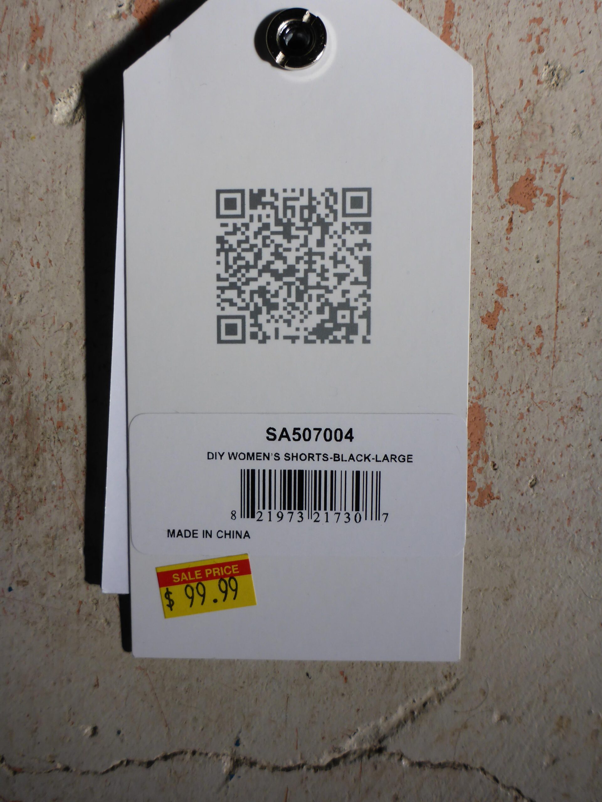 Race Face DIY shorts: Alt text: A clothing tag displaying a QR code, product code "SA507004," and description "DIY Women&rsquo;s Shorts - Black - Large." The tag features a sale price of $99.99 and indicates that the item is made in China. The background shows a textured wall.