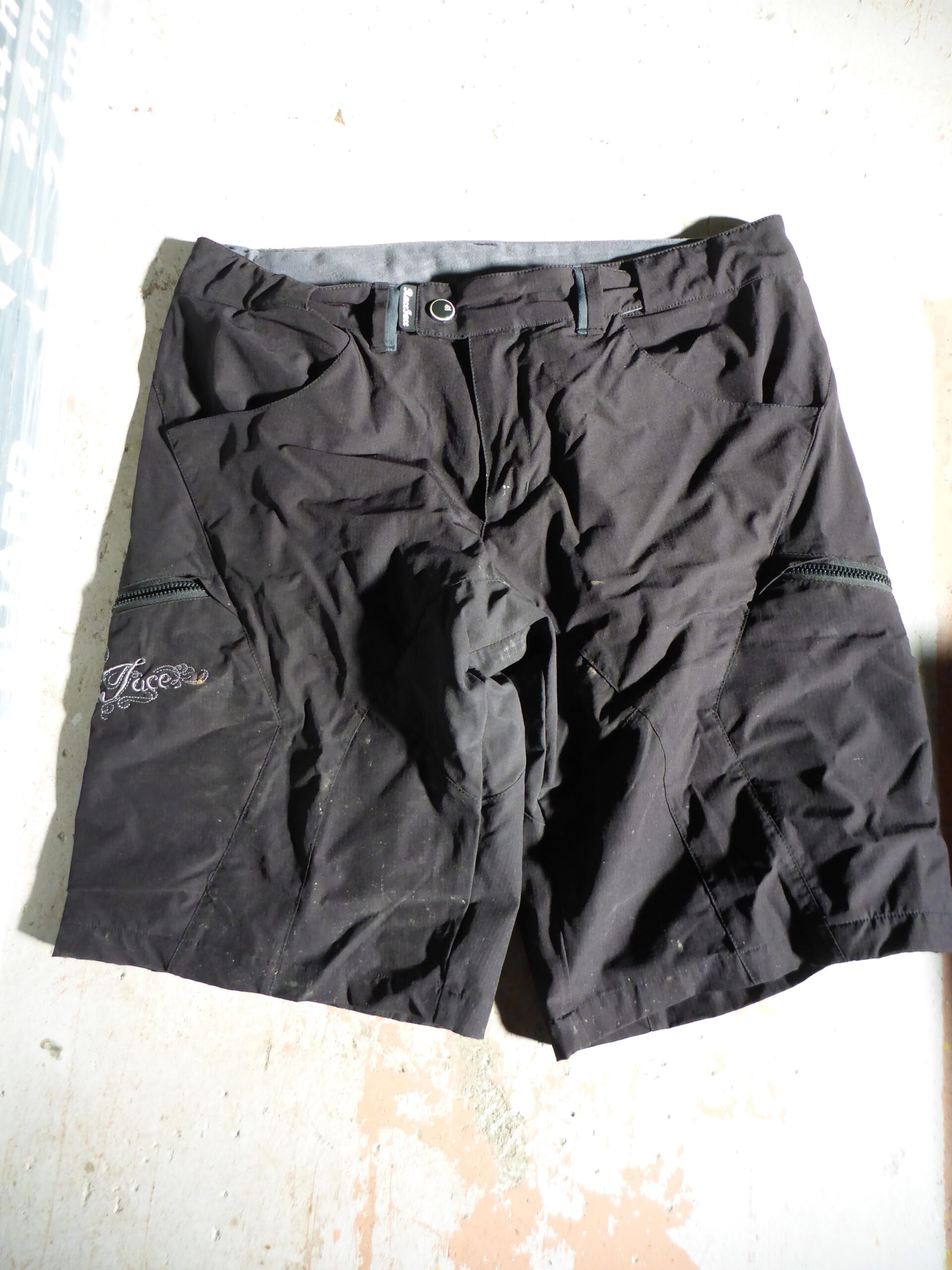 Race Face Piper shorts: Black outdoor shorts laid flat on a surface, featuring side pockets with zippers and a waistband with a button and belt loops. The fabric appears slightly wrinkled and dusty.