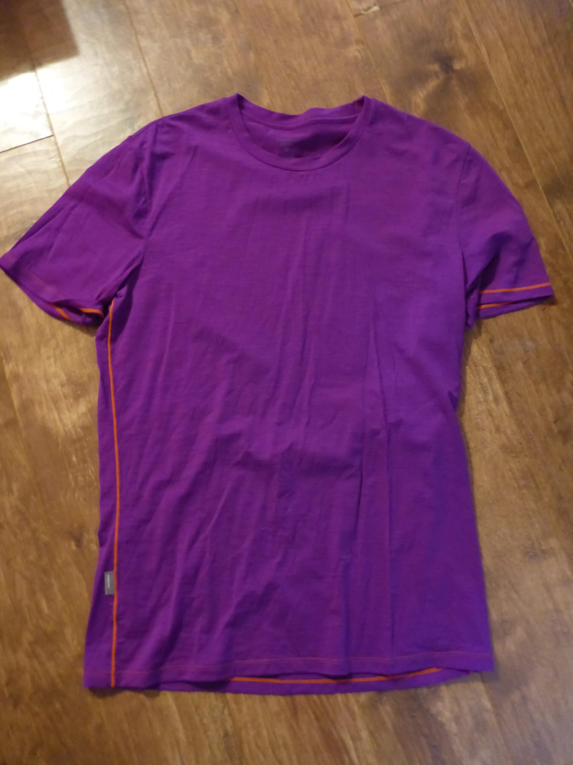 Icebreaker t-shirt: A folded purple T-shirt with short sleeves, featuring orange stitching along the sleeves and hem, laid out flat on a wooden surface.