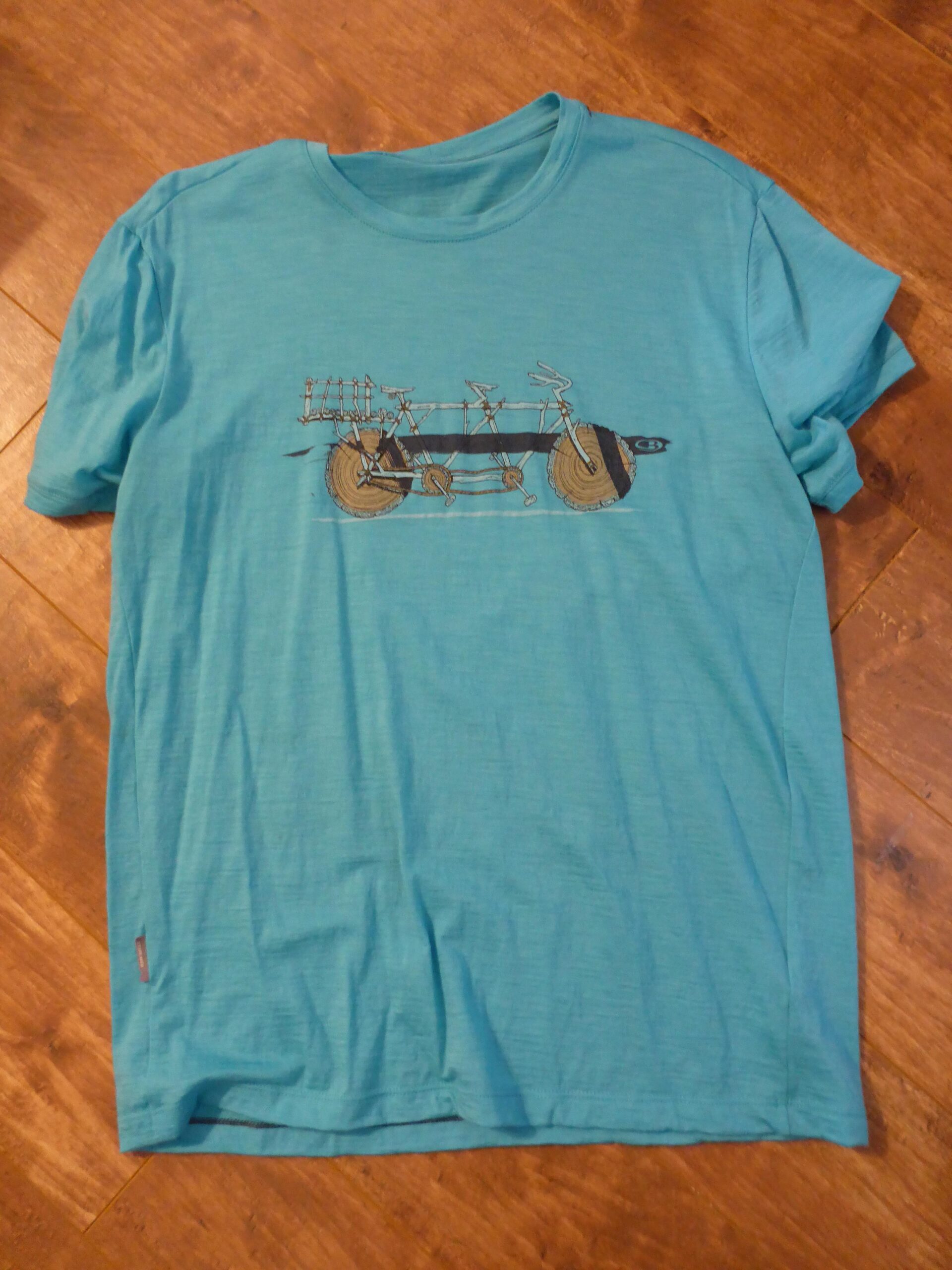 Icebreaker t-shirt: A turquoise t-shirt laid flat on a wooden surface, featuring a graphic design of a bicycle with unusual wheels and handlebars.
