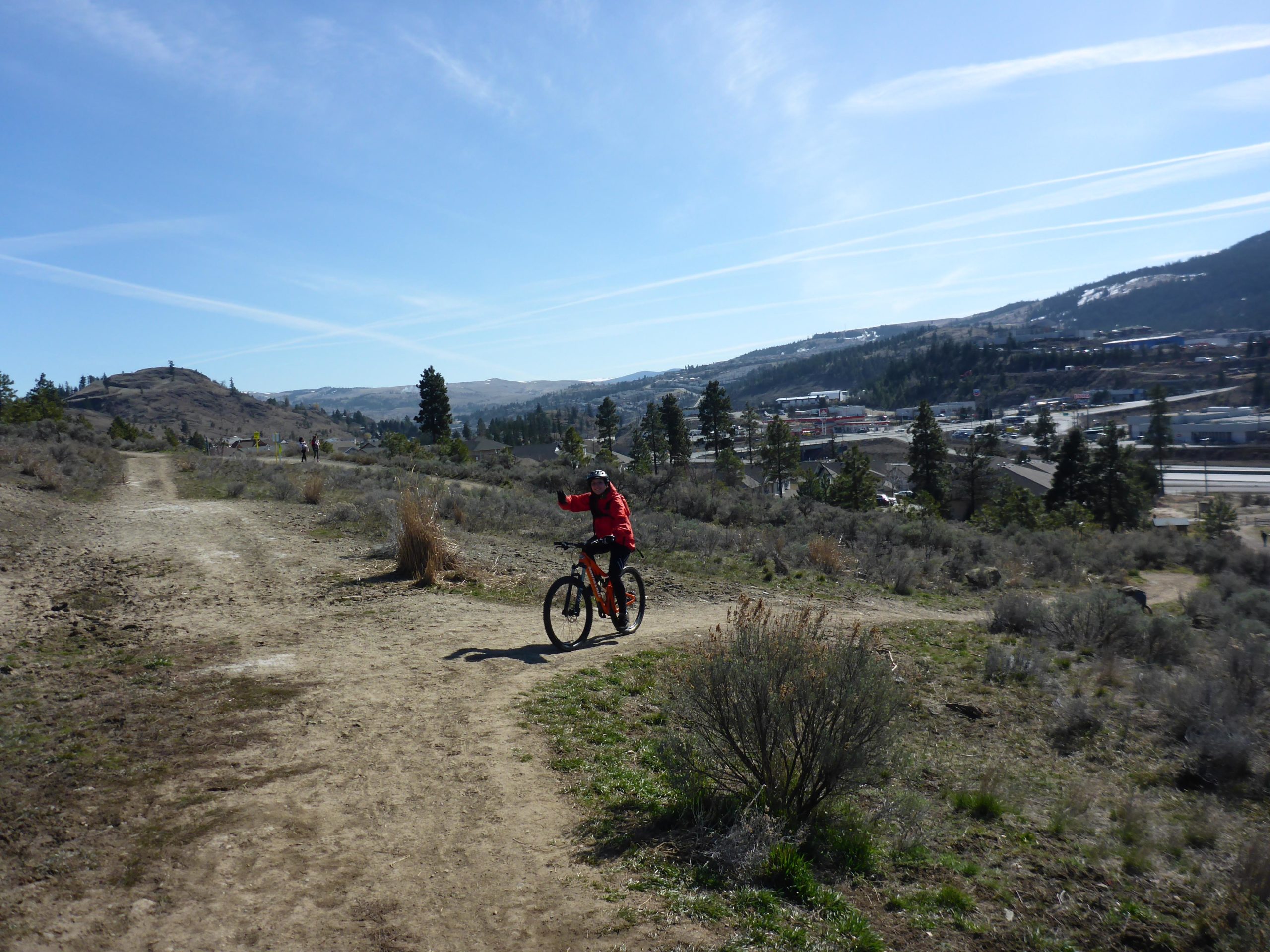 A person in a red jacket riding a mountain bike along a dirt trail in a hilly outdoor area. The background includes distant hills, trees, and a cityscape, with clear blue skies and wispy clouds. Kenna Cartwright Park mountain bike trail.