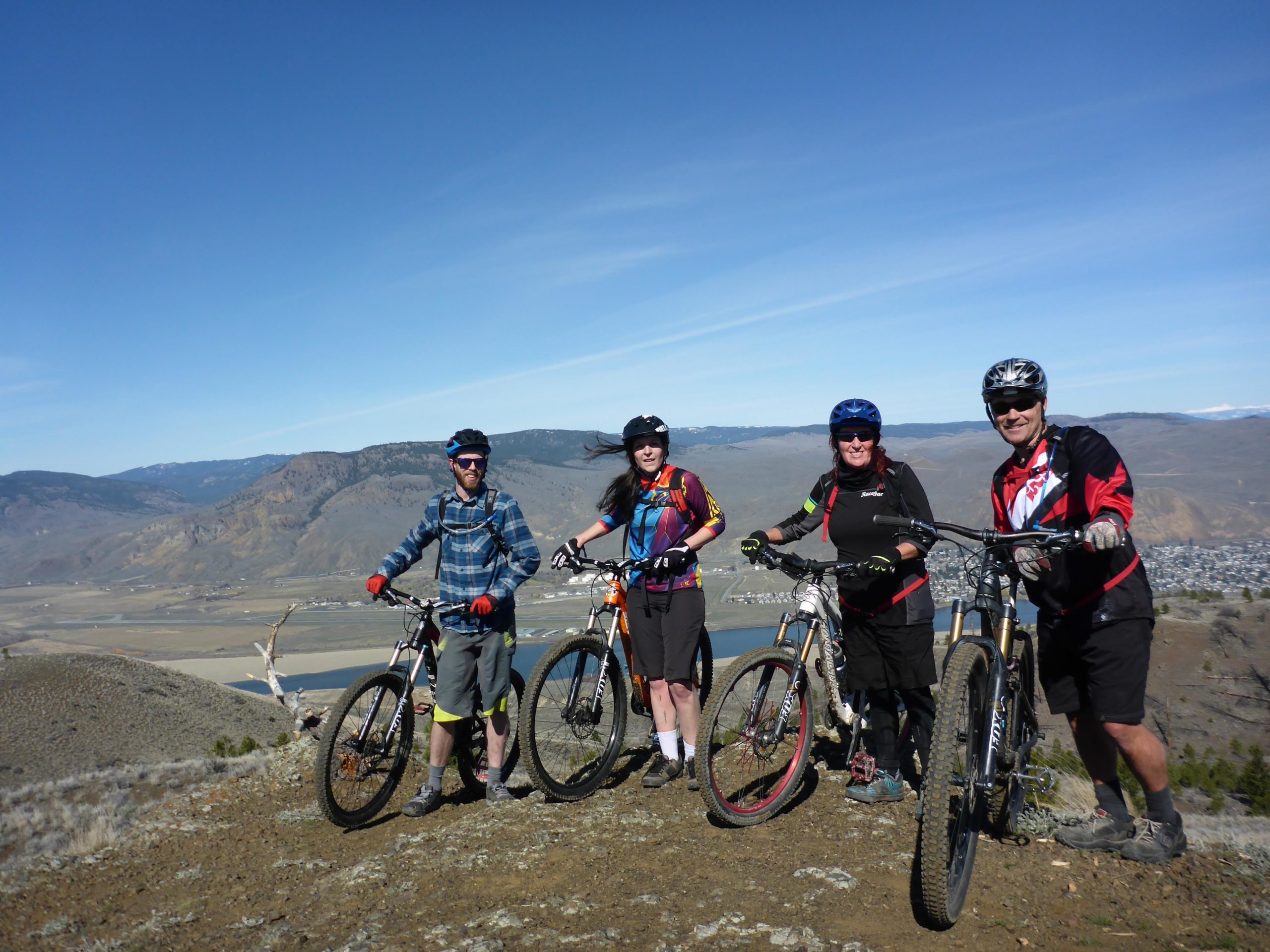 Four mountain bikers posing at a scenic overlook, with rolling hills and a valley in the background. They are wearing helmets and cycling gear, and each is standing next to their mountain bikes. The sky is clear and blue, suggesting a sunny day. Kenna Cartwright Park mountain bike trail.