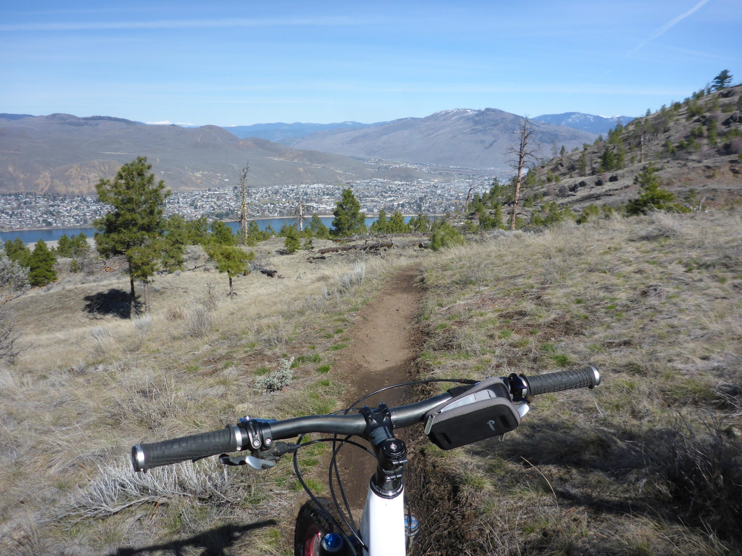 A mountain biking view from the handlebars, showing a dirt trail leading down a hillside with scattered trees. In the background, a lake and a small town are visible, framed by mountains under a clear blue sky. Kenna Cartwright Park mountain bike trail.