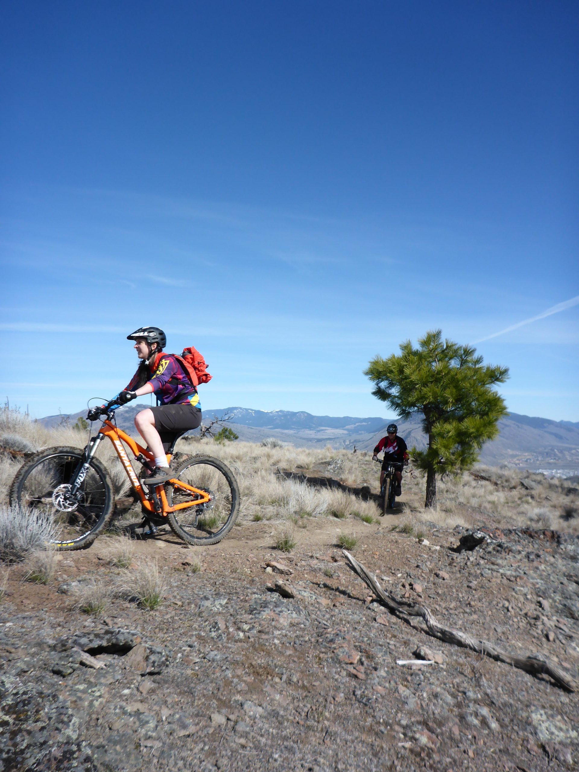 A person riding a mountain bike on a rocky trail in a natural landscape. The rider is wearing a helmet and a colorful shirt, and an orange backpack is visible. In the background, another mountain biker is approaching, with a green tree and mountains visible under a clear blue sky. Kenna Cartwright Park mountain bike trail.