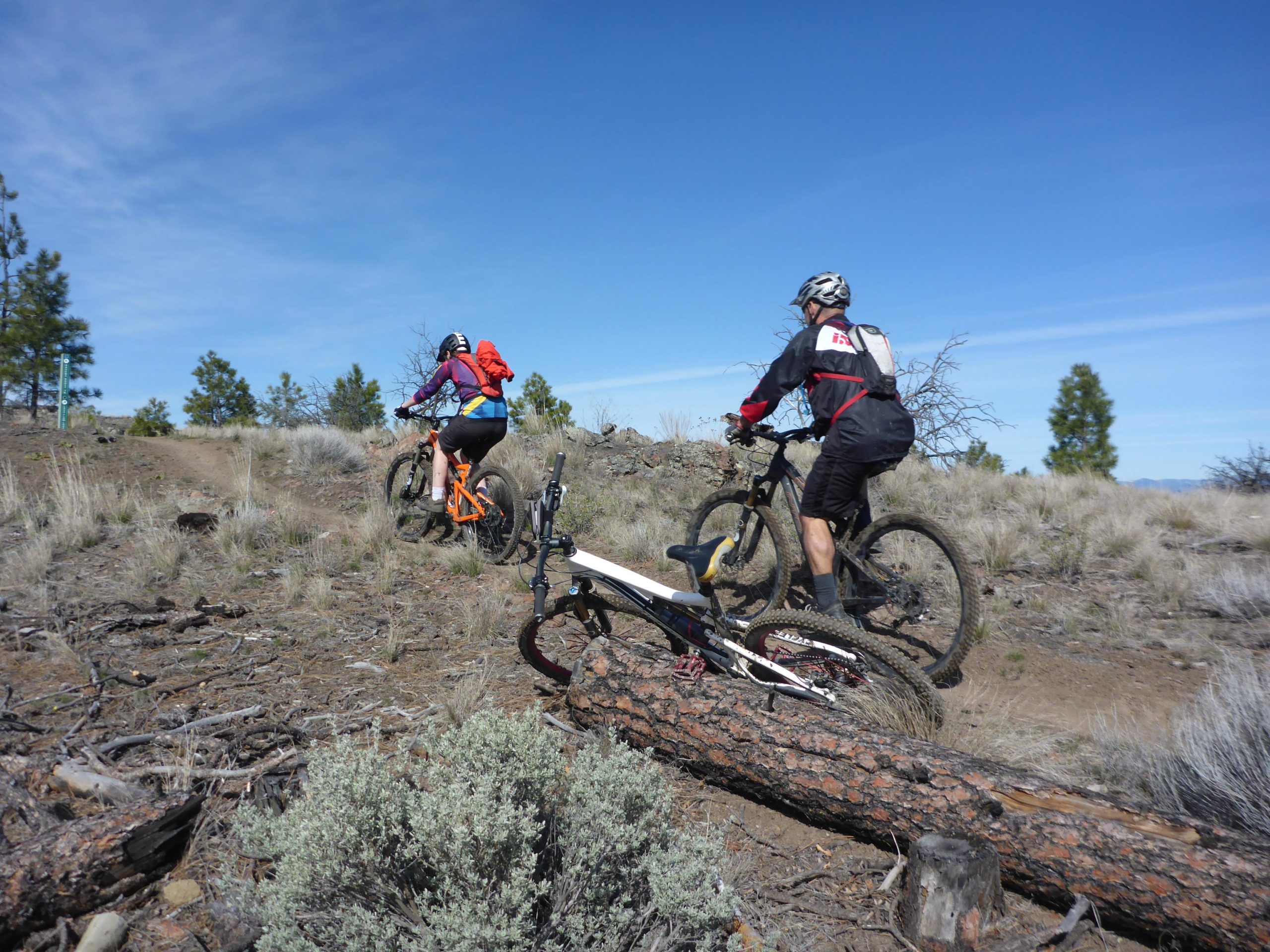 Two mountain bikers navigate a rugged trail in a rocky, open landscape. One rider, wearing an orange bike and a red backpack, is ahead, while the second rider, dressed in black and red gear, follows closely behind. In the foreground, a bike lies on its side next to a fallen log, with scattered vegetation in the dry terrain. The sky above is clear and blue, indicating a sunny day. Kenna Cartwright Park mountain bike trail.