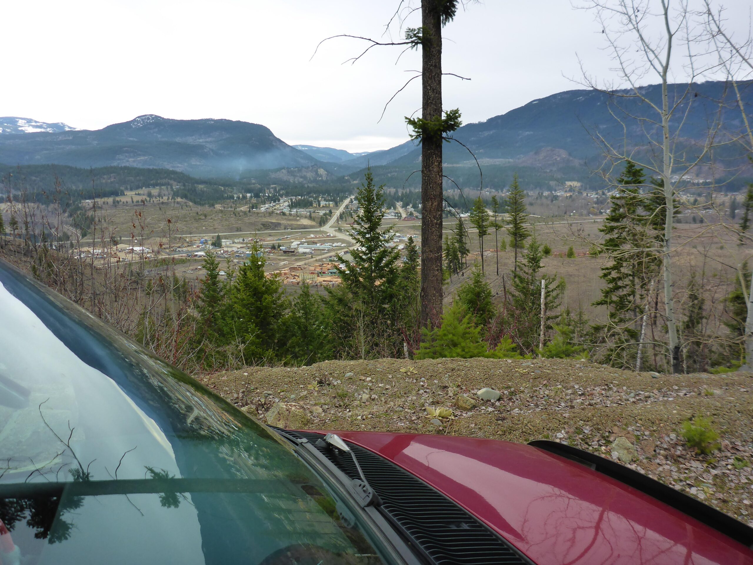 A view of a valley from a hillside, featuring a red vehicle's windshield in the foreground. Dense greenery, including trees, is visible on both sides, while a small town can be seen in the distance, nestled within rolling hills under a cloudy sky. Bluegrouse Ridge Trail mountain bike trail.