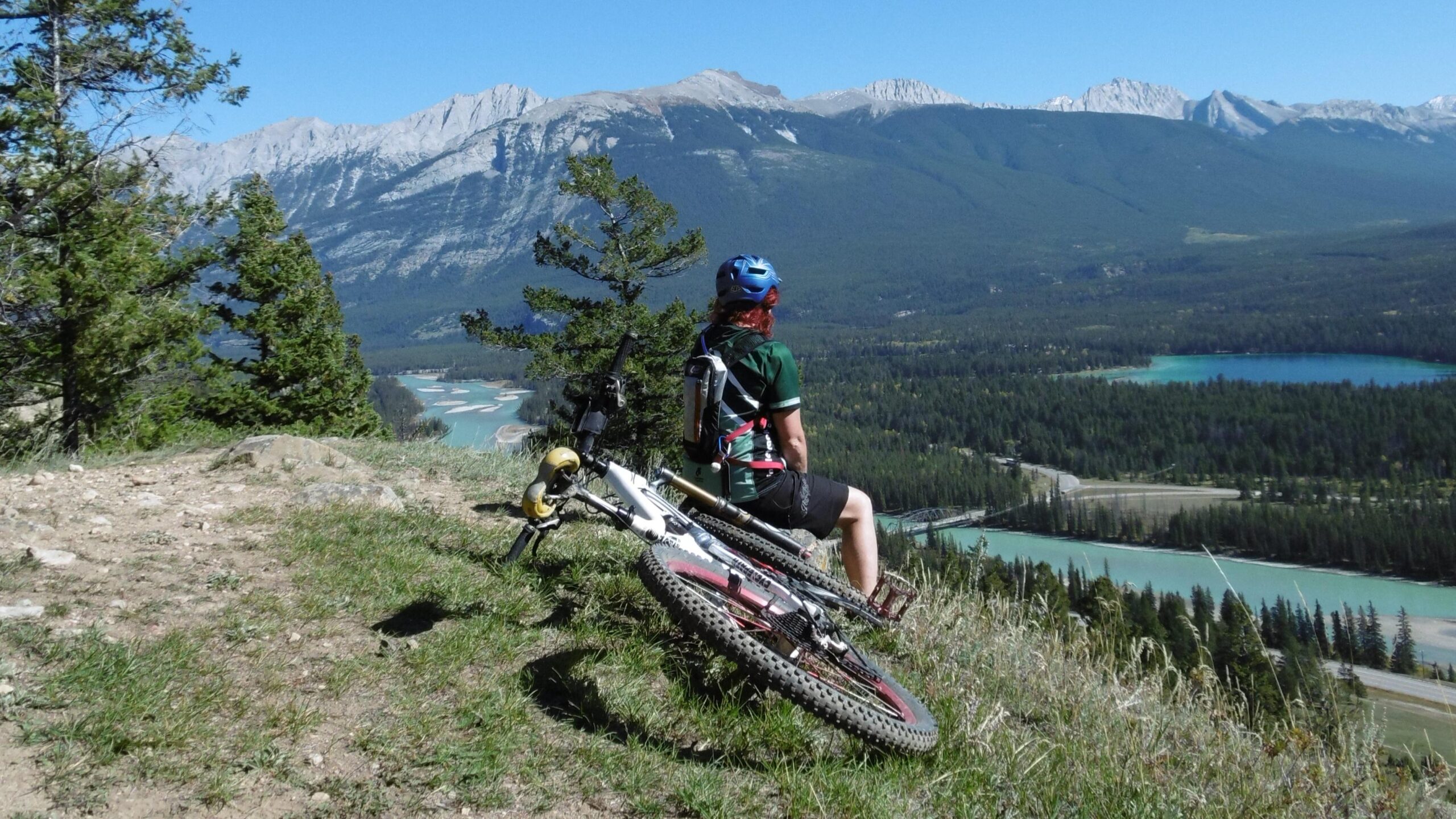 A mountain biker sits on a grassy hill overlooking a scenic landscape. In the background, there are majestic mountains and a winding river surrounded by trees. The biker, wearing a helmet and a green jersey, rests next to their bicycle, enjoying the view on a sunny day. Pyramid Bench mountain bike trail.