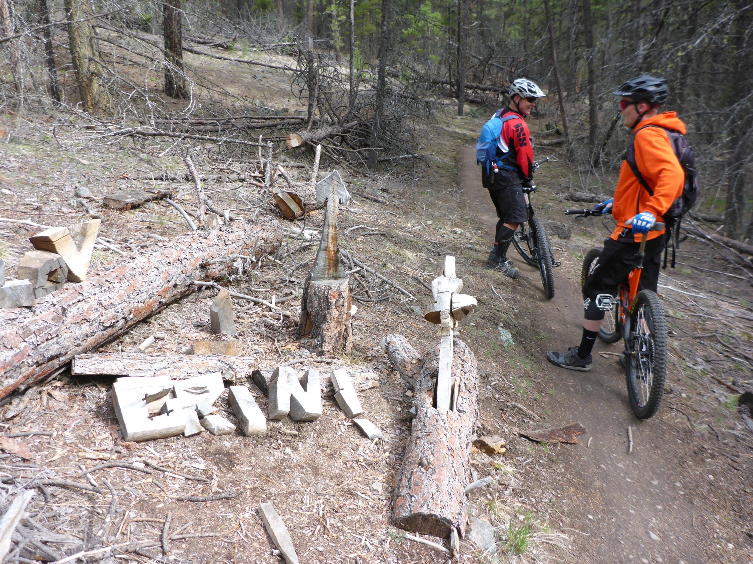 Two mountain bikers pause on a forest trail surrounded by trees, with one wearing an orange hoodie and the other in a red and blue jersey. Nearby, carved pieces of wood spell out "ERIN!" on the ground, and a wooden sign points down the trail. Smith Creek mountain bike trail.