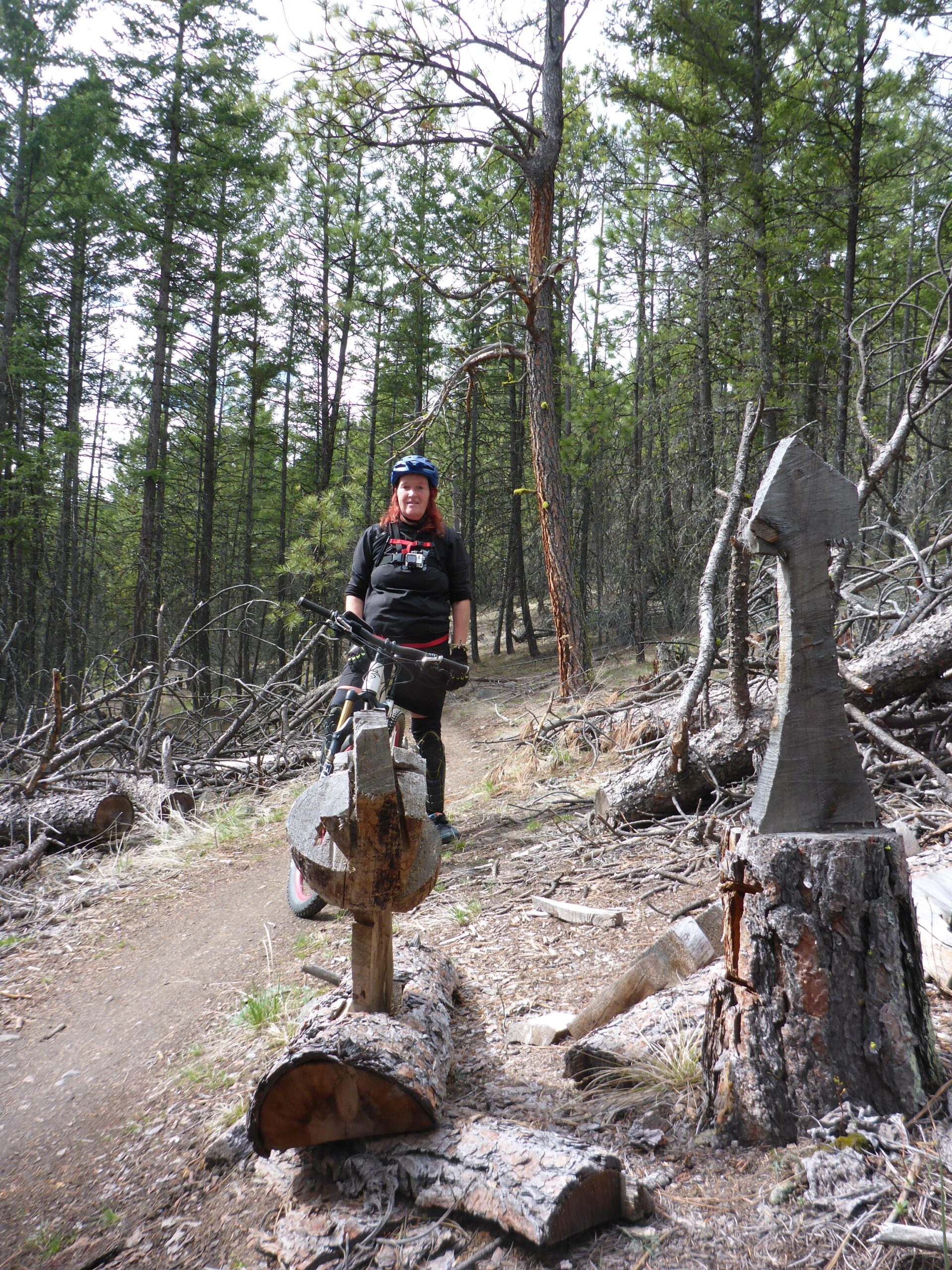 A person in a cycling helmet stands next to a wooden trail marker on a narrow path through a forest. The background features tall trees and scattered logs, suggesting a natural outdoor setting suitable for mountain biking. Smith Creek mountain bike trail.