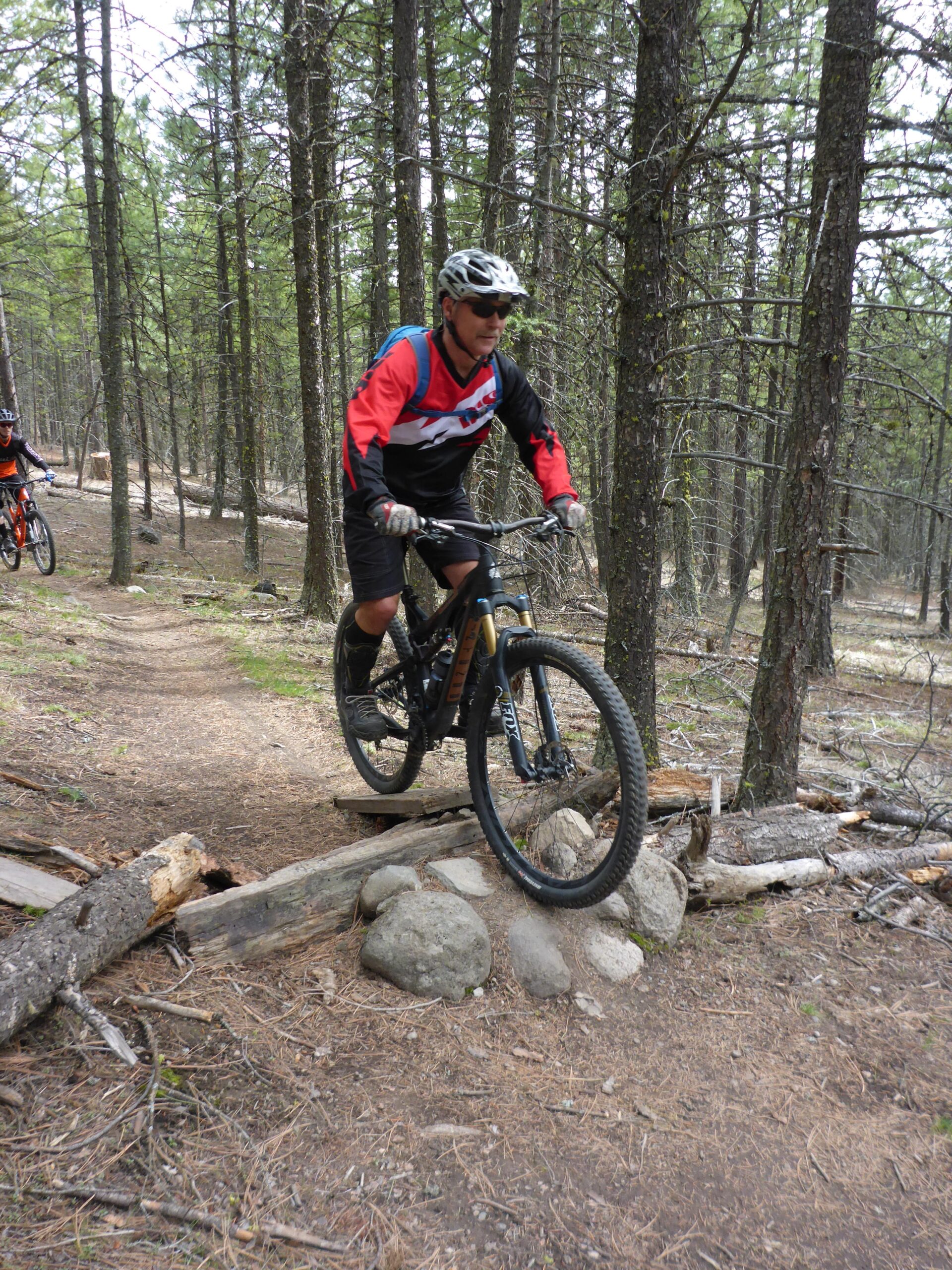 A mountain biker navigating a rocky section of a wooded trail, with trees in the background and a second biker visible in the distance. The rider is wearing a helmet and sunglasses, and has a focused expression as he rides over a small wooden bridge supported by rocks. Smith Creek mountain bike trail.