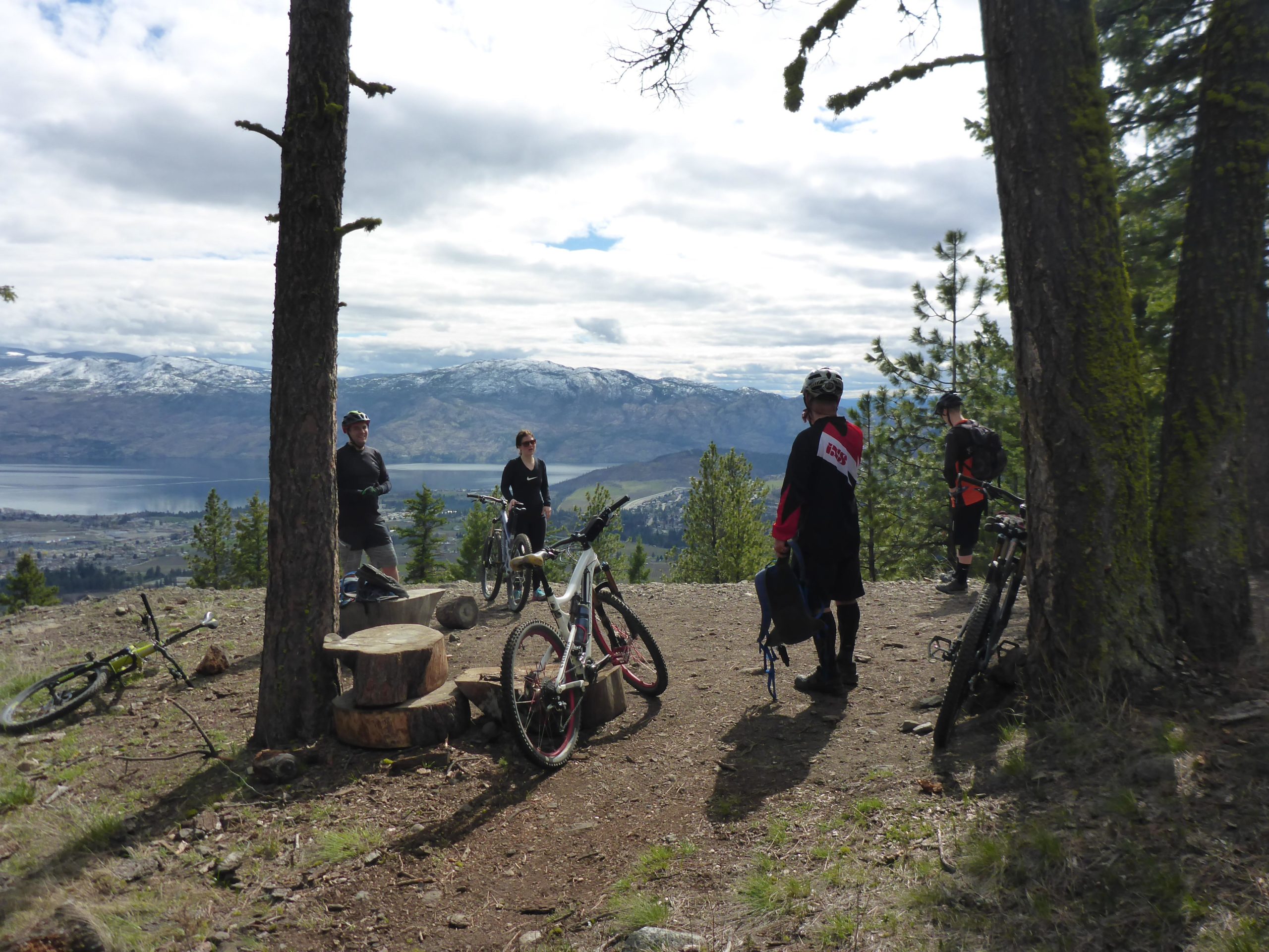 Four mountain bikers standing near their bikes at a viewpoint overlooking a lake and mountainous landscape. The scene is set amidst trees, with snow-capped mountains in the background and a cloudy sky above. Some bikers are conversing while others are in a relaxed pose, enjoying the view. Smith Creek mountain bike trail.