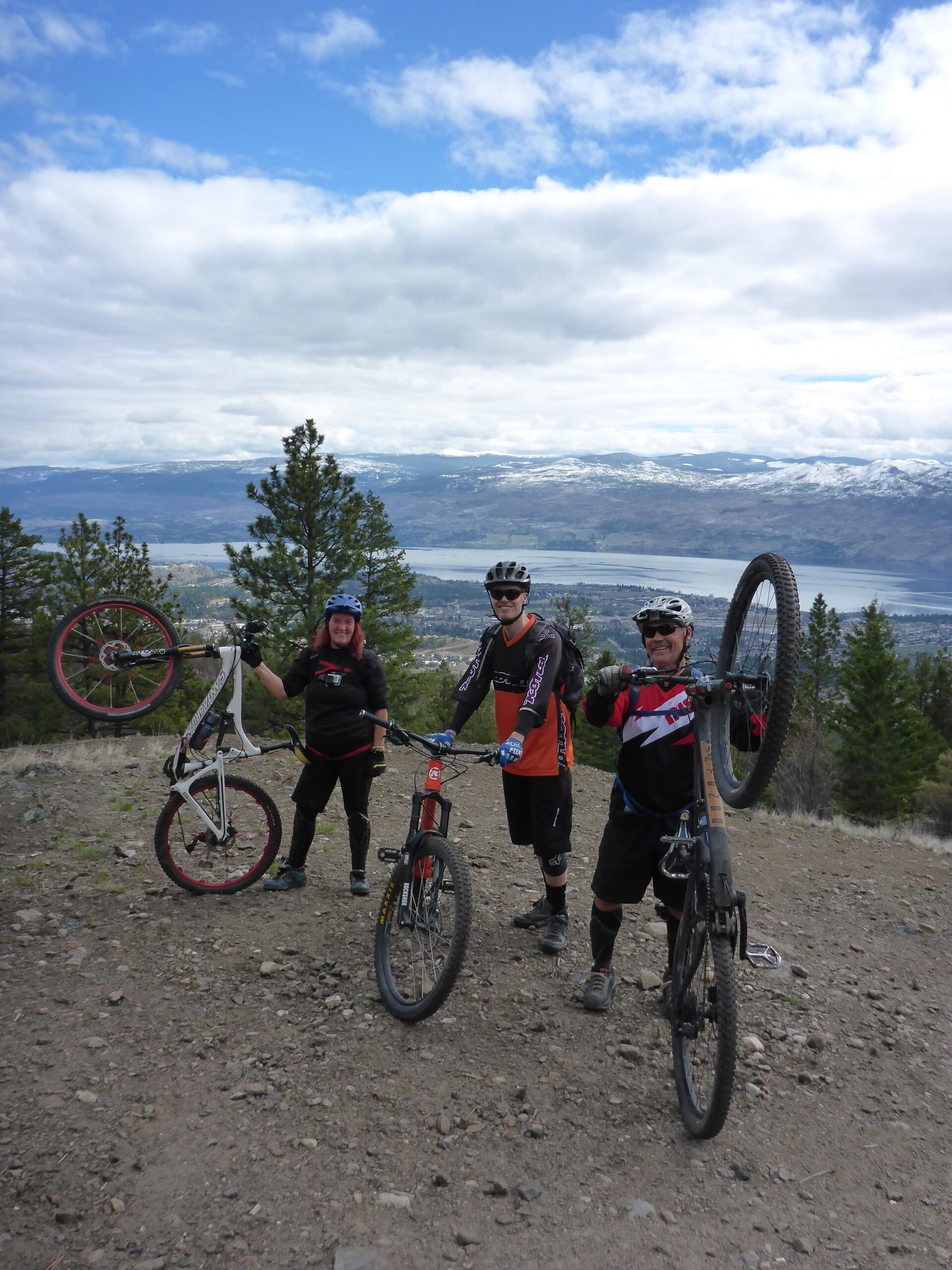 Three mountain bikers pose on a rocky trail overlooking a scenic valley. They are dressed in cycling gear, with two of them holding their bikes in a playful manner—one raising a bike above their head, while another holds a bike beside them. Lush greenery and distant mountains are visible under a partly cloudy sky. Smith Creek mountain bike trail.