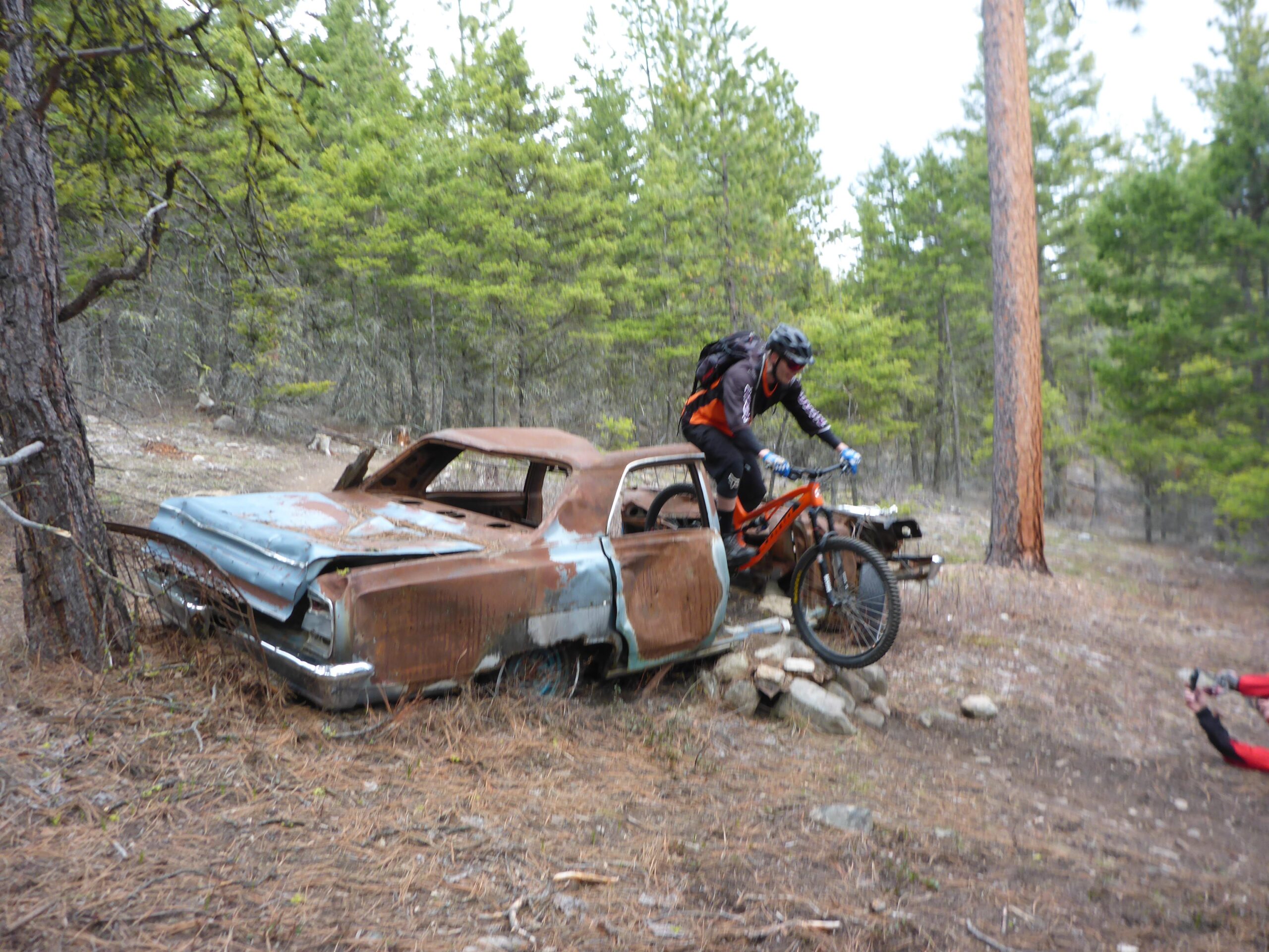 A mountain biker in a black helmet and orange and black gear performs a trick over the roof of an abandoned, rusted car surrounded by trees and pine needles in a forested area. Smith Creek mountain bike trail.