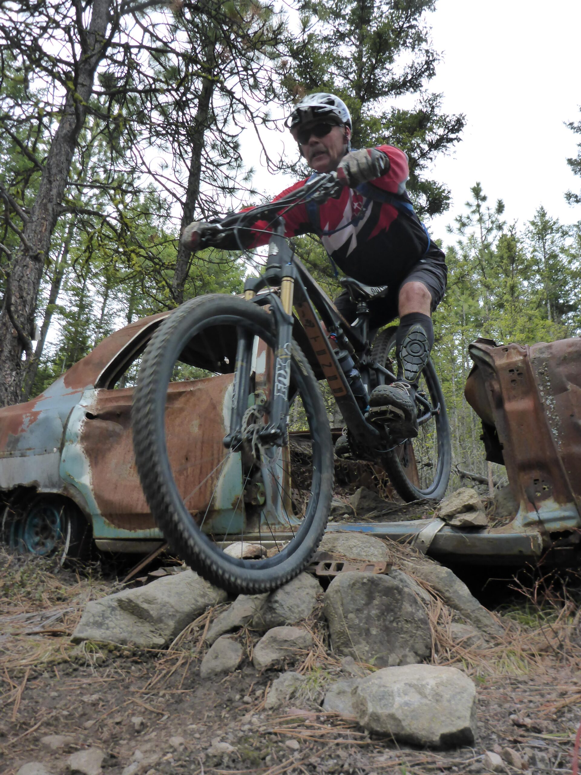 A mountain biker in a red and black jersey rides over rocky terrain, navigating between the remains of rusted, abandoned cars in a forested area. Trees and greenery surround the scene, creating a natural backdrop. Smith Creek mountain bike trail.