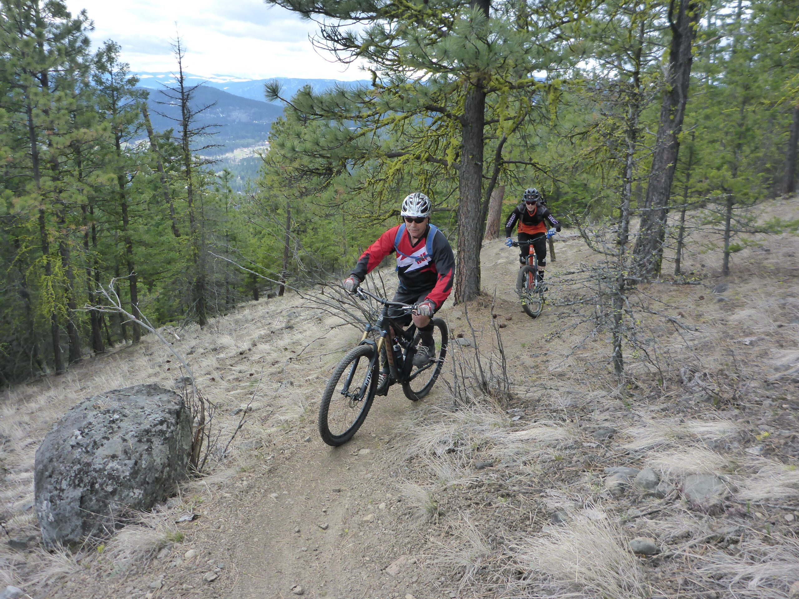 Two mountain bikers navigate a winding trail through a forested area with tall green trees and rocky terrain. One biker is in the foreground wearing a red and black jersey, while the second biker, further back, is in a black and orange outfit. The scene captures the rugged outdoor environment and the thrill of mountain biking. Smith Creek mountain bike trail.