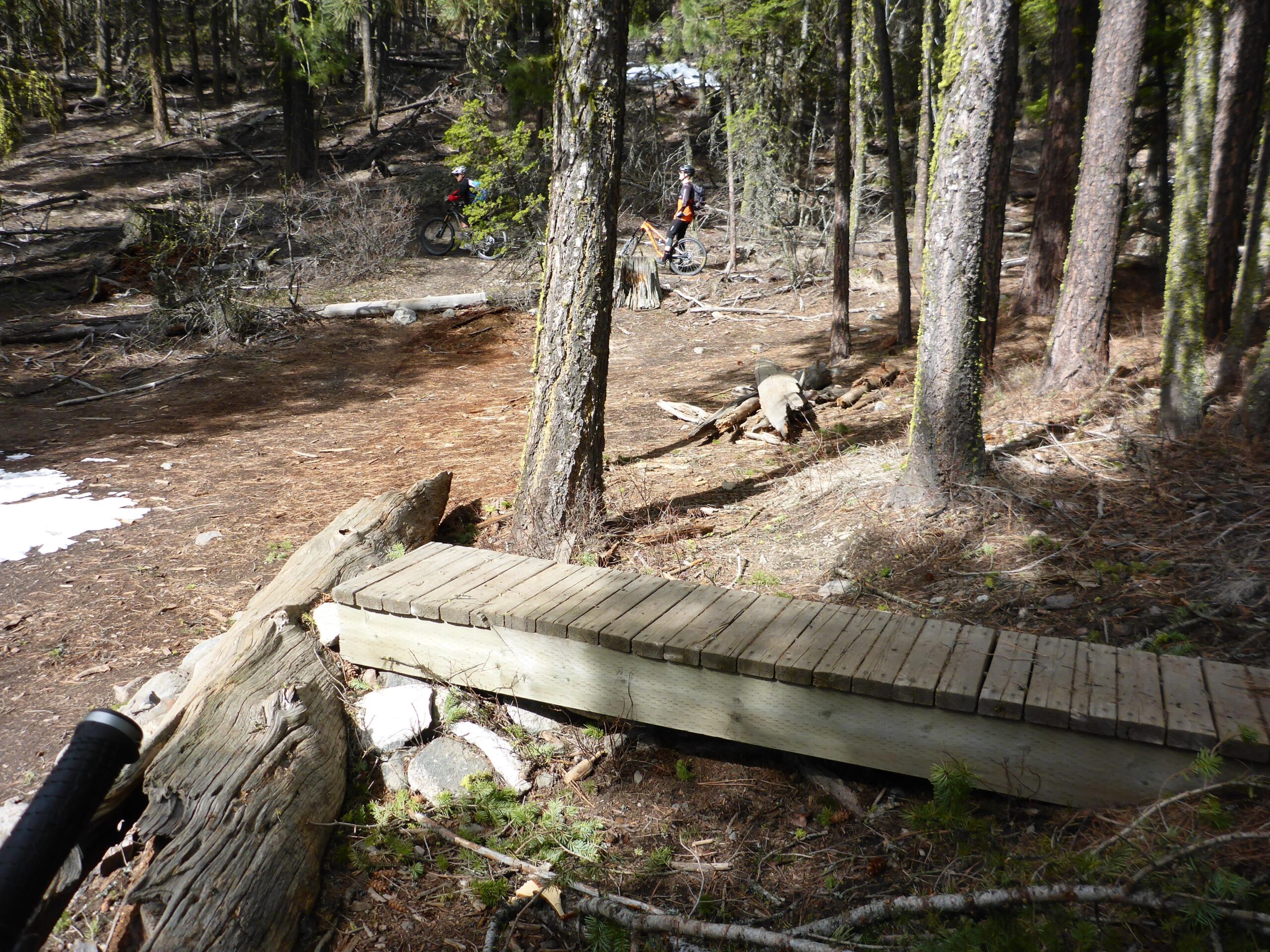 A wooden bridge over a small area of forest floor covered in pine needles and debris, leading through a wooded area with tall trees. In the background, a person is seen riding a bicycle on a trail. There are patches of snow on the ground nearby. Smith Creek mountain bike trail.