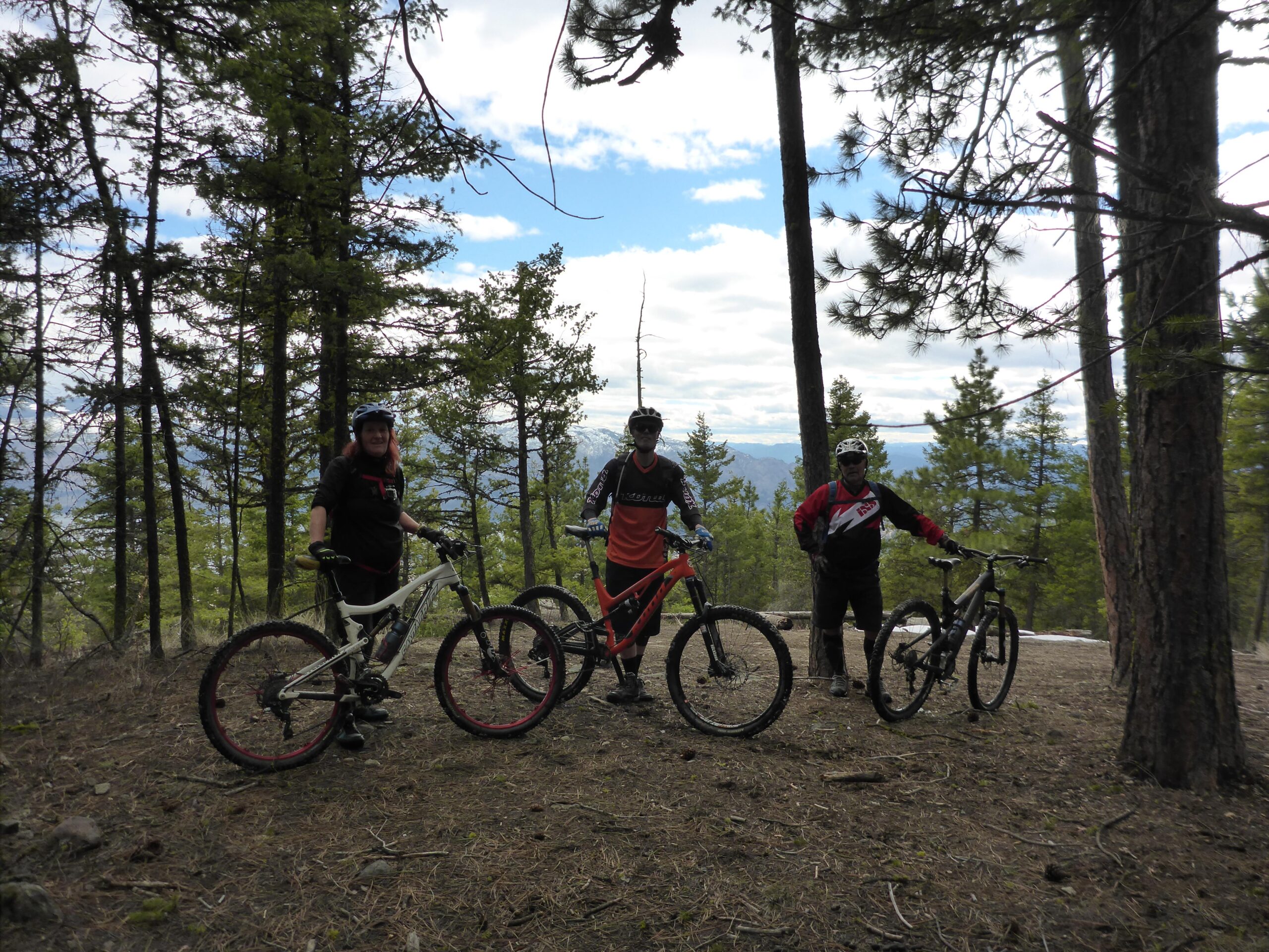 Three mountain bikers pose with their bikes in a forested area. They are surrounded by tall pine trees and a cloudy sky. One rider has reddish-brown hair and is wearing a helmet and gloves, standing beside a white bike. The second rider is dressed in red and black, standing next to an orange mountain bike. The third rider is wearing a black and white outfit and is next to a dark bike, with distant mountains visible in the background. Smith Creek mountain bike trail.